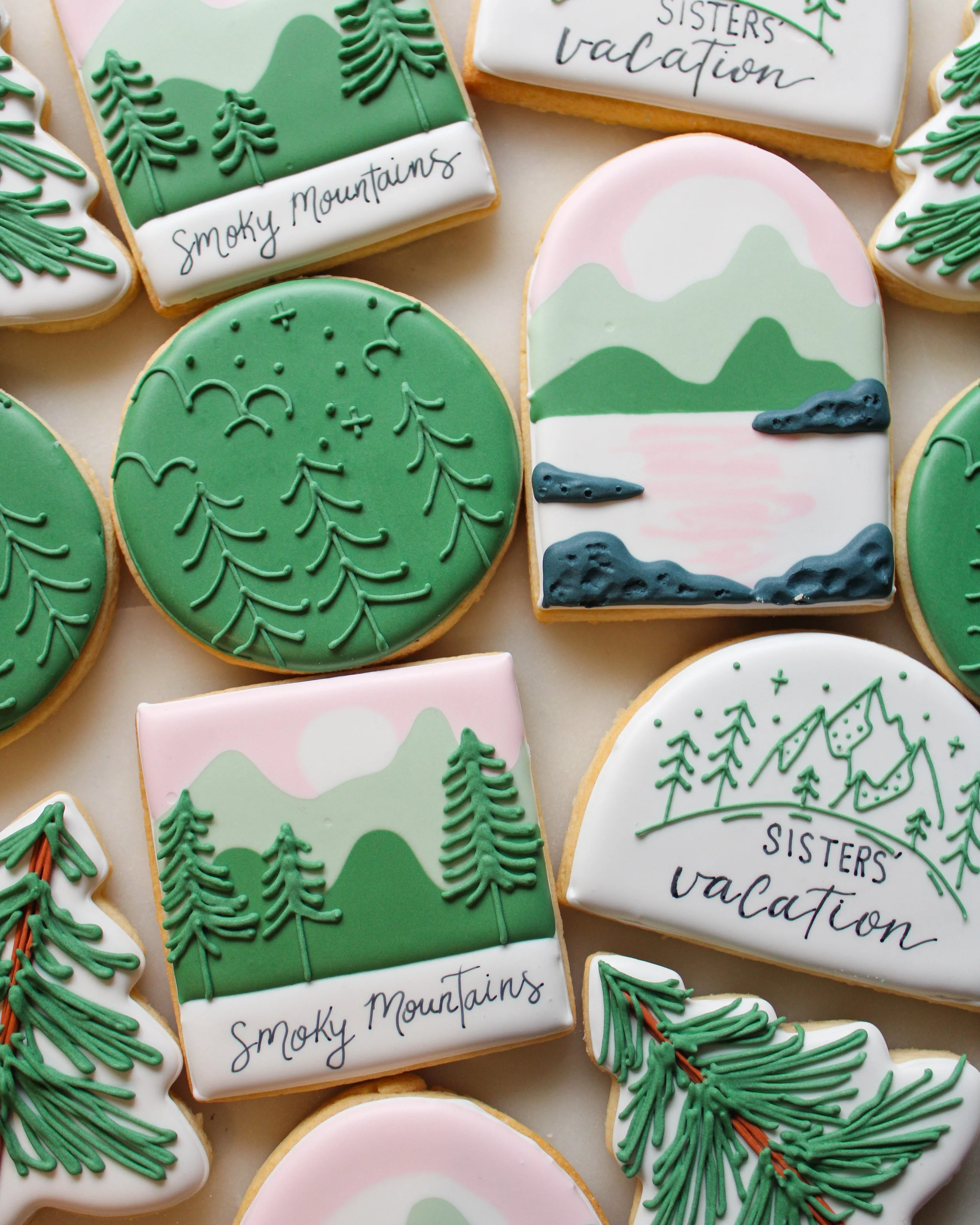 Decorative cookies with green pine trees, mountains, and sunset designs, some with text like 'Smoky Mountains' and 'Sisters vacation'.