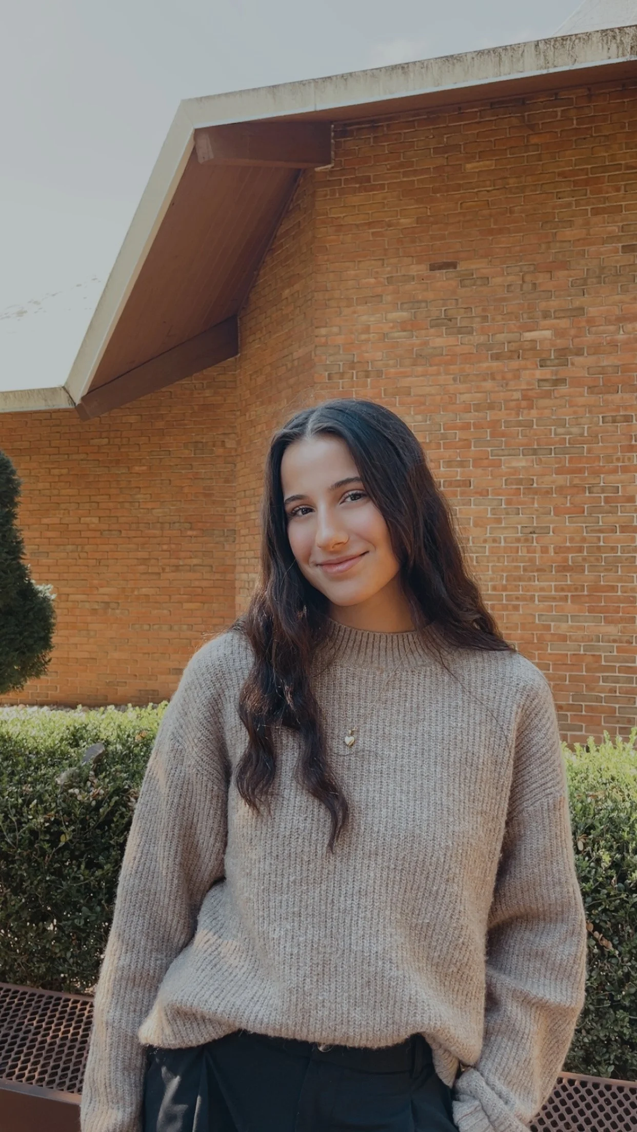 A young woman with long dark hair wearing a beige sweater and black pants standing outdoors in front of a brick building with a manicured hedge and a bench.