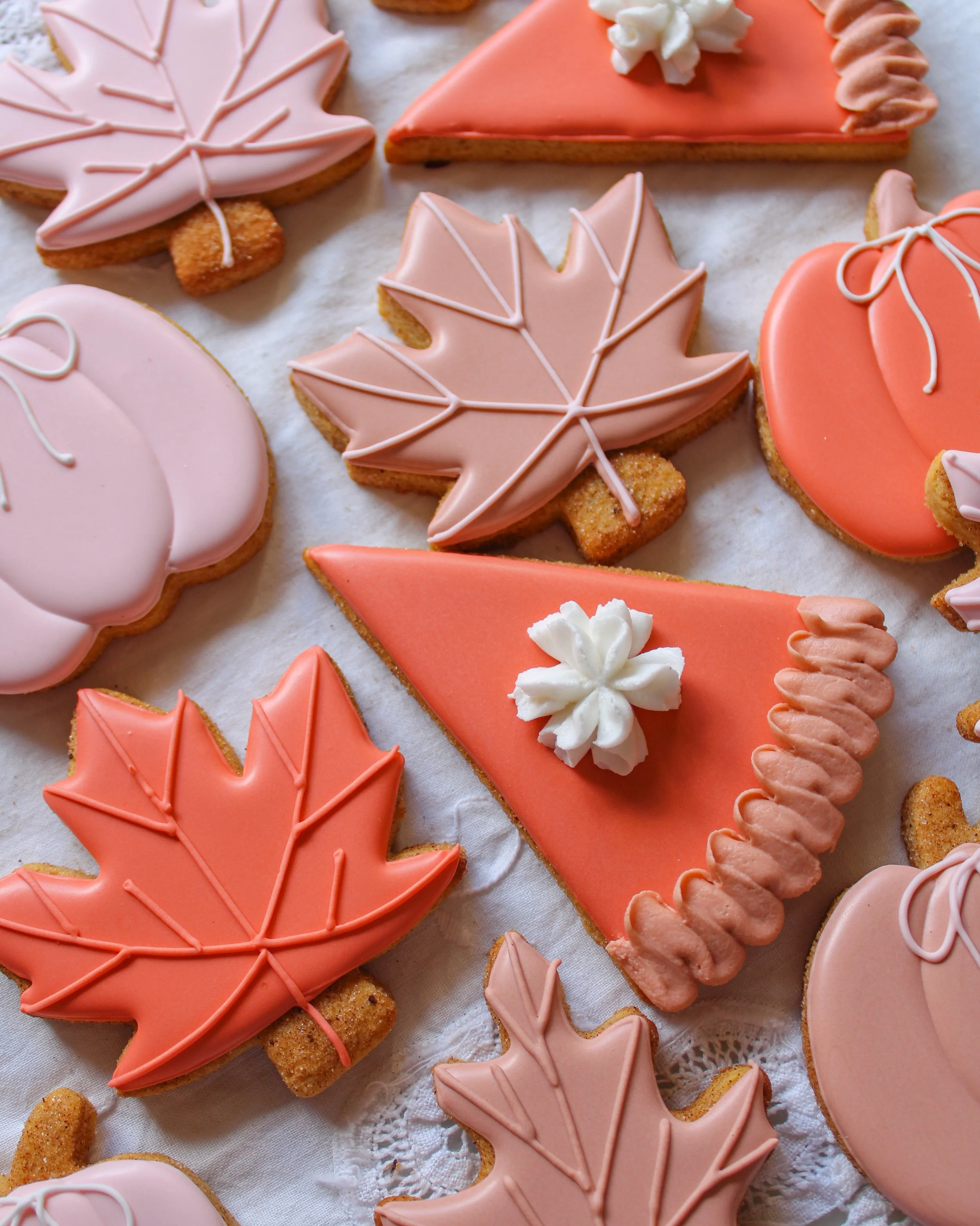 Assorted autumn-themed cookies decorated with pink and orange icing, shaped like leaves, pumpkins, and slices of pumpkin pie.