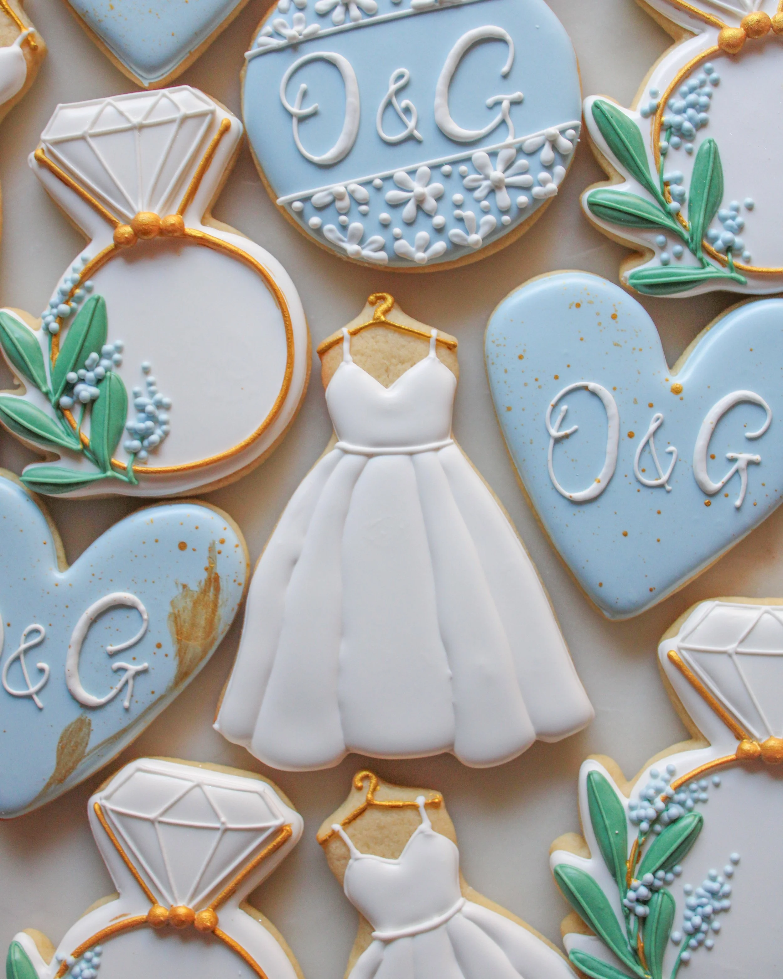 Decorated cookies with wedding theme, including a white wedding dress, rings, and hearts with personalized initials 'O & G', decorated with pastel blue, white, and gold icing and floral accents.