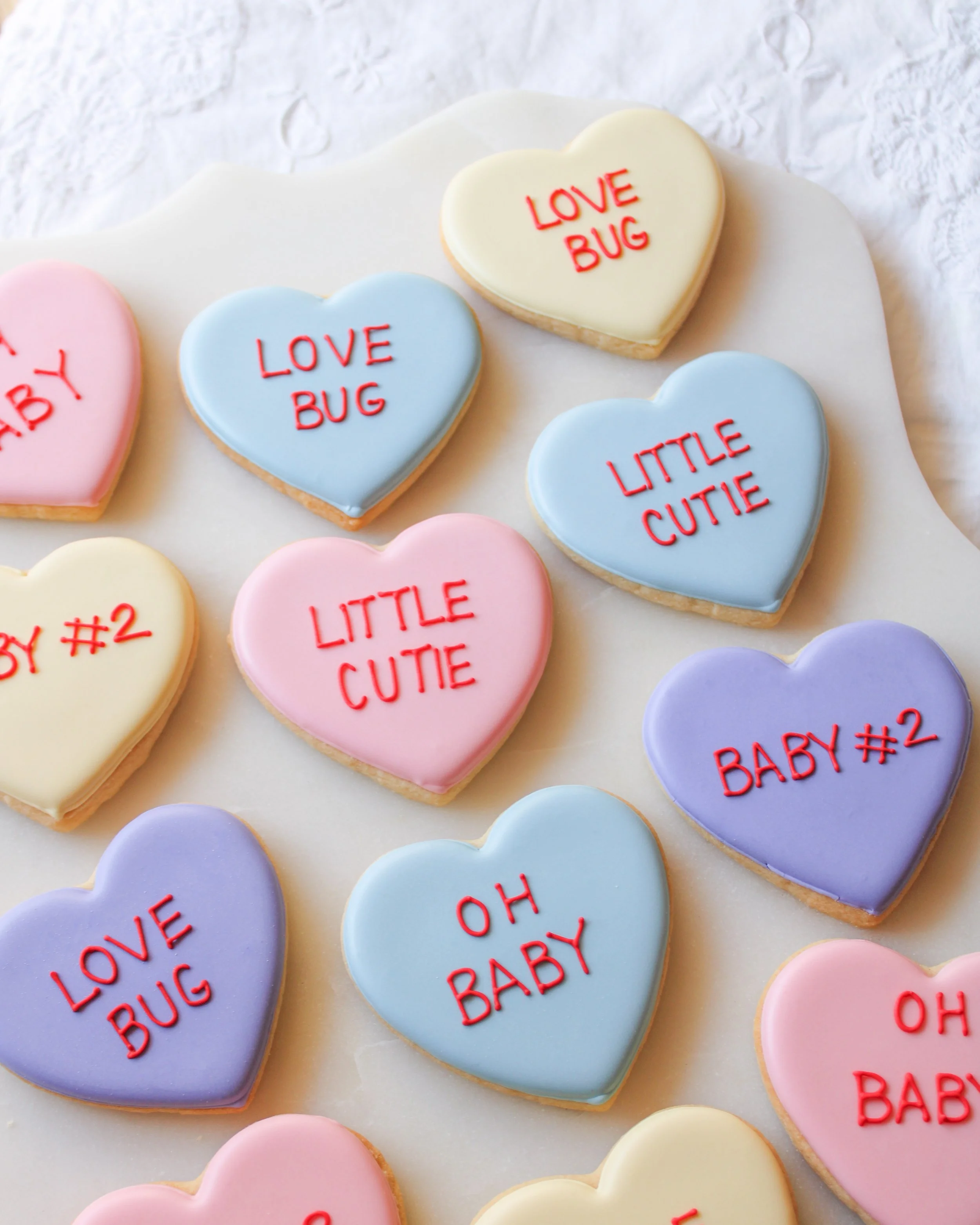 conversation heart baby announcement cookies decorated with pastel-colored icing and red writing, featuring phrases like 'LOVE BUG,' 'LITTLE CUTIE,' 'BABY #2,' and 'OH BABY,' arranged on a white surface.