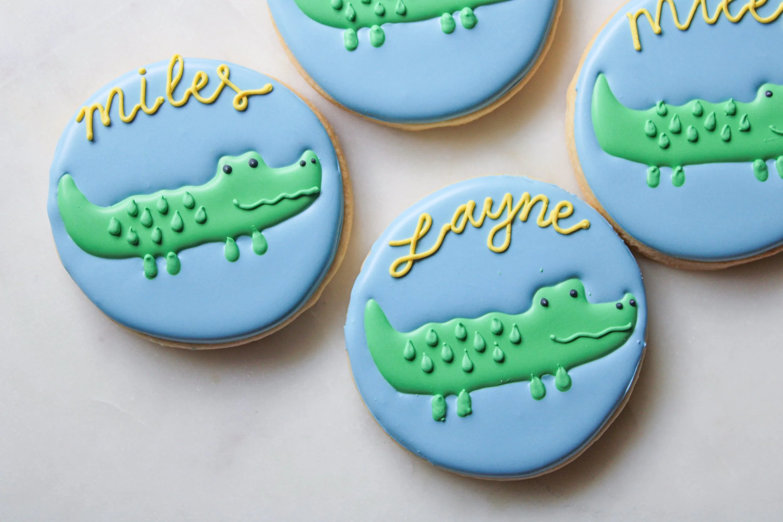 Decorative cookies featuring a green crocodile on a blue background, with the names 'Miles' and 'Layne' written in yellow icing.