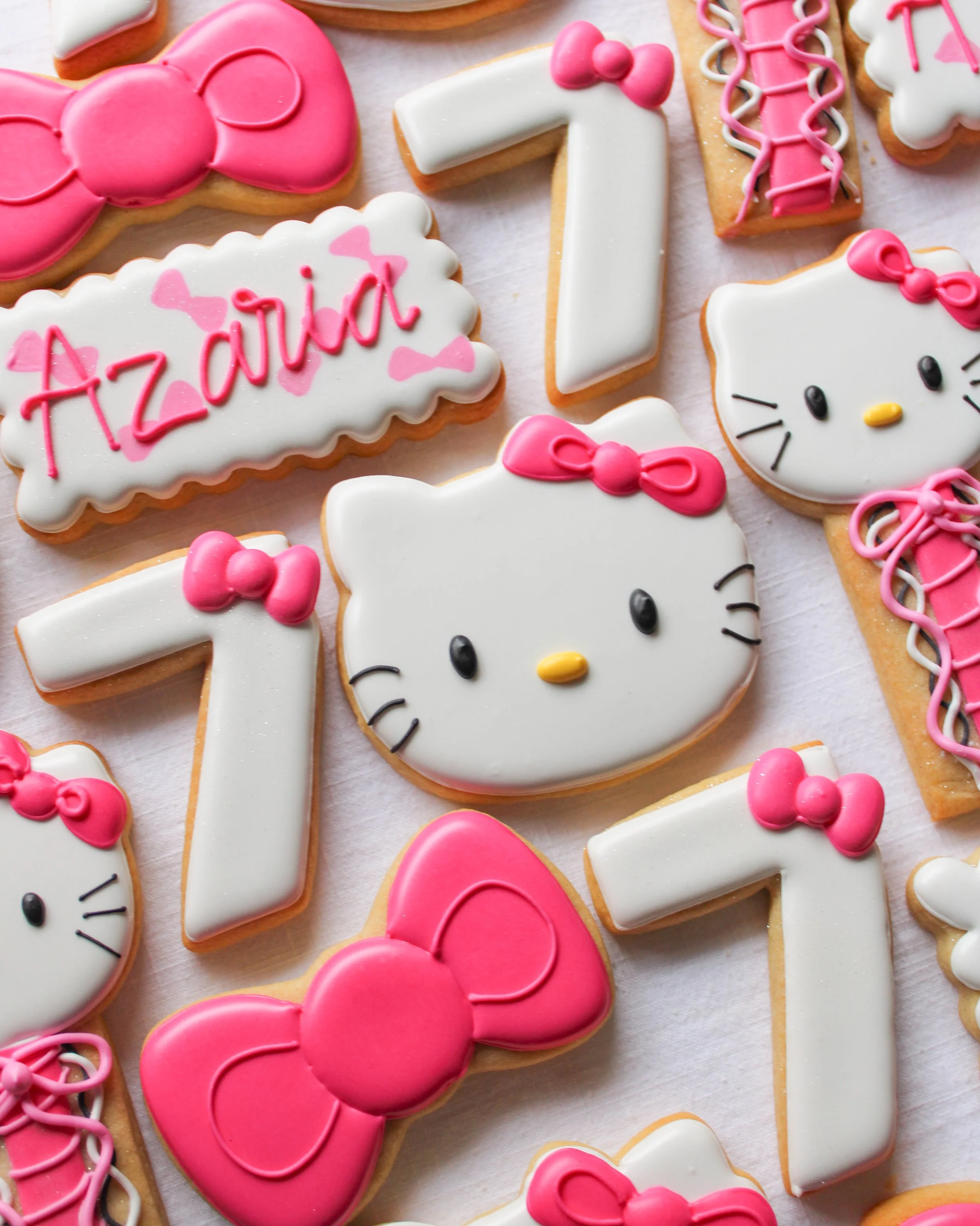 Decorated sugar cookies with Hello Kitty faces, pink bows, the number seven, and a cookie with the name "Azaria" in pink icing, set on a white surface.