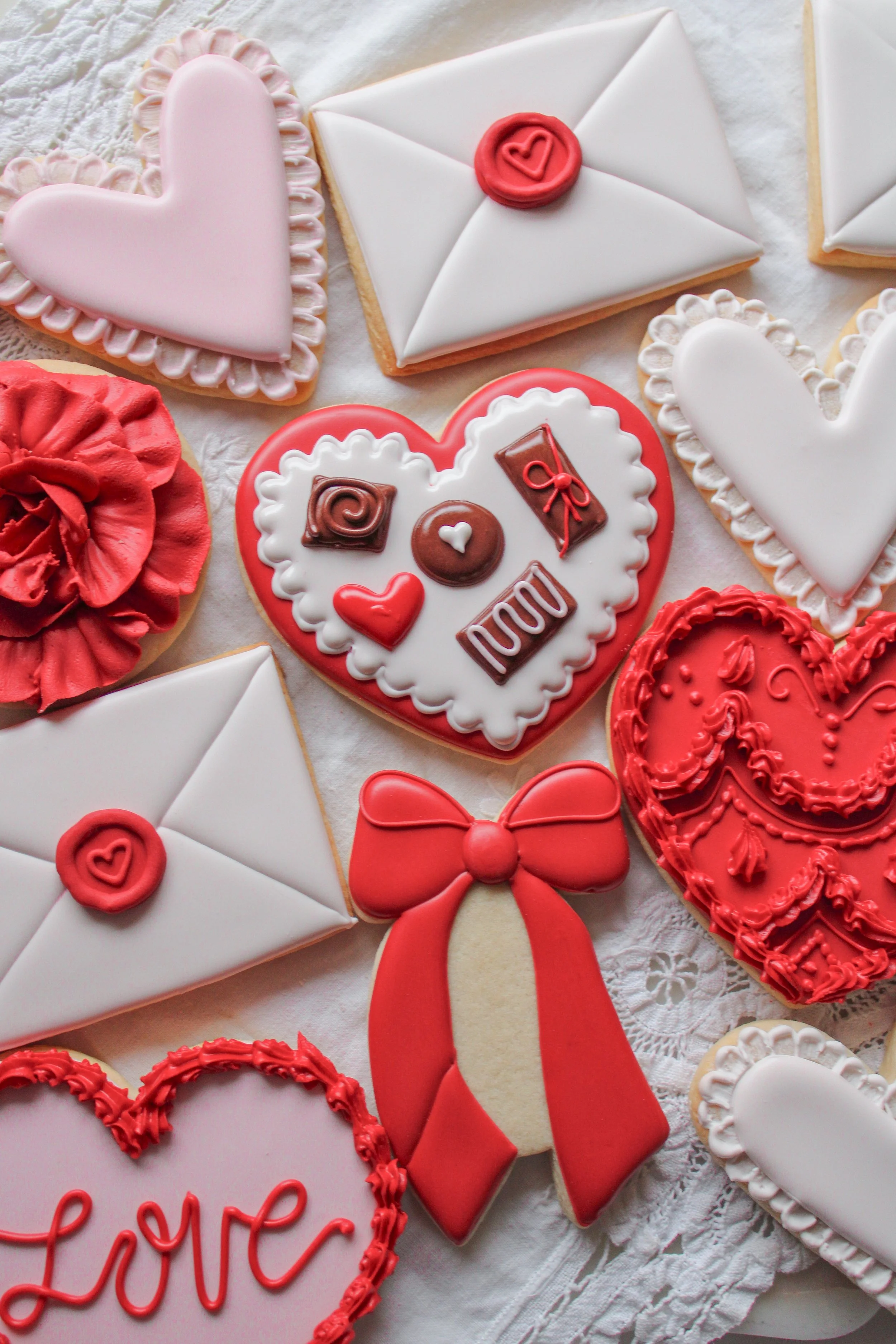 Assorted Valentine's Day cookies decorated with pink, red, and white icing, including hearts, envelopes, chocolates, a bow, and the word 'Love' written on one cookie.