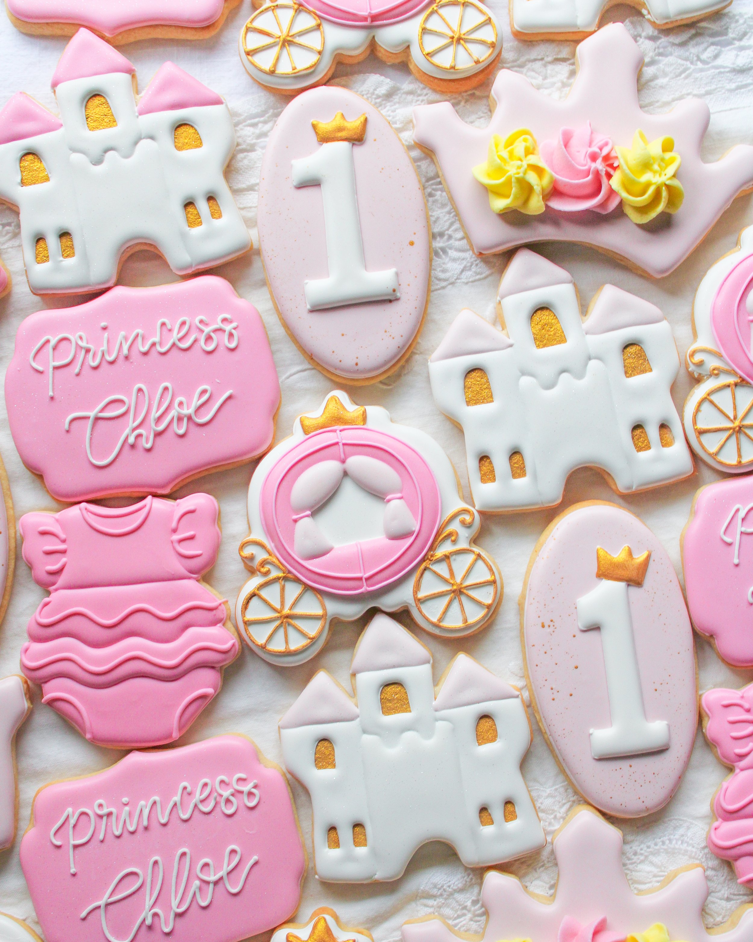 Decorated princess-themed cookies in pink, white, and yellow with castles, a princess crown, a carriage, and the number 1 for a first birthday celebration.