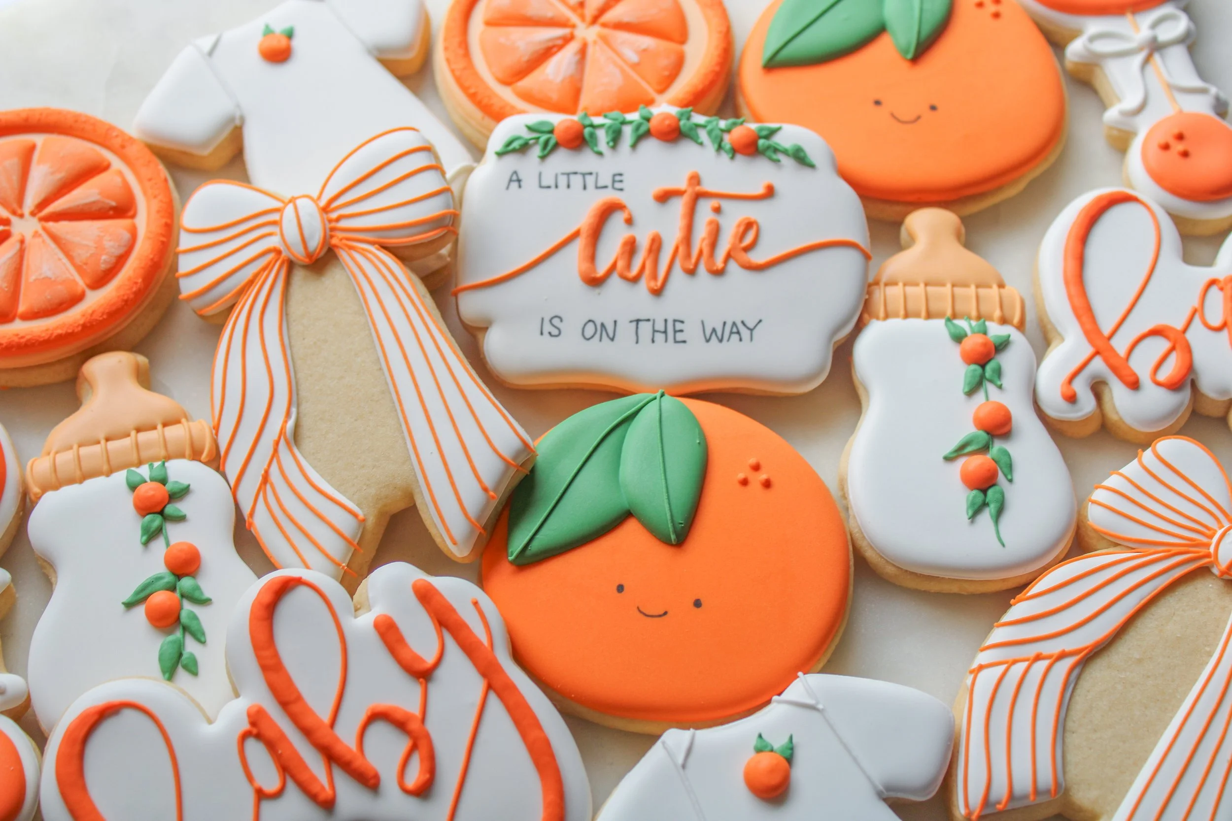 Decorative baby shower sugar cookies themed with oranges, baby bottles, and a message that says 'A little cutie is on the way.' The cookies are decorated with orange, green, and white icing, featuring smiling faces, leaves, and striped bows.