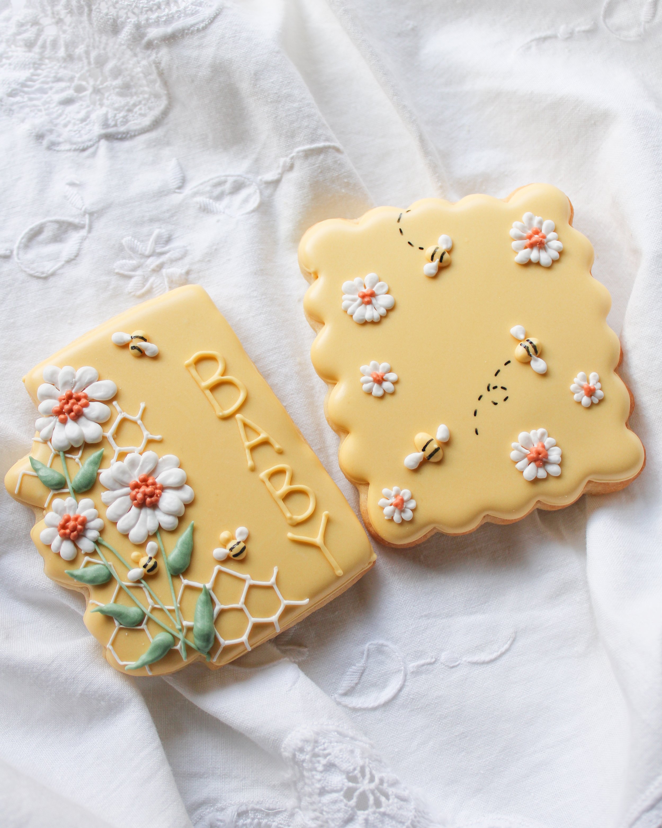 Decorated yellow cookies with bees, flowers, and honeycomb patterns, one with the word 'BABY' written on it, on white embroidered fabric.