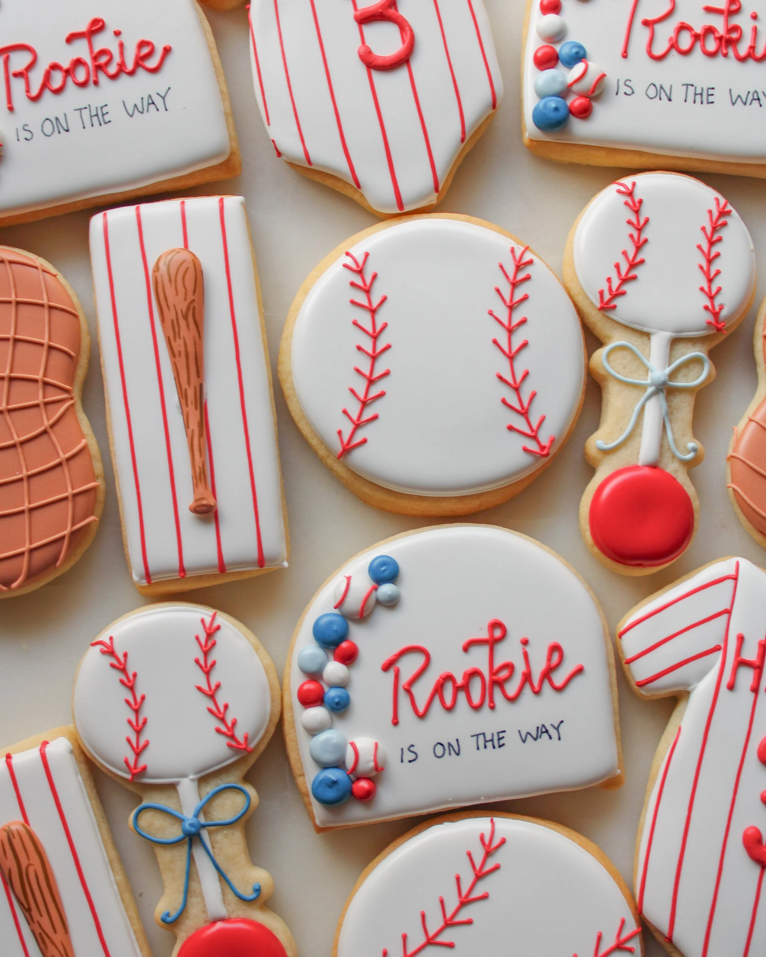 Decorated baseball-themed cookies, including cookies shaped like baseballs, baseball bats, and jerseys, with red, white, and blue icing and writing that says "Rookie is on the way."