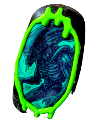 A cake pop with an image of an xenomorph inspired by the film Alien, surrounded by a neon glow in the dark green chocolate drip.