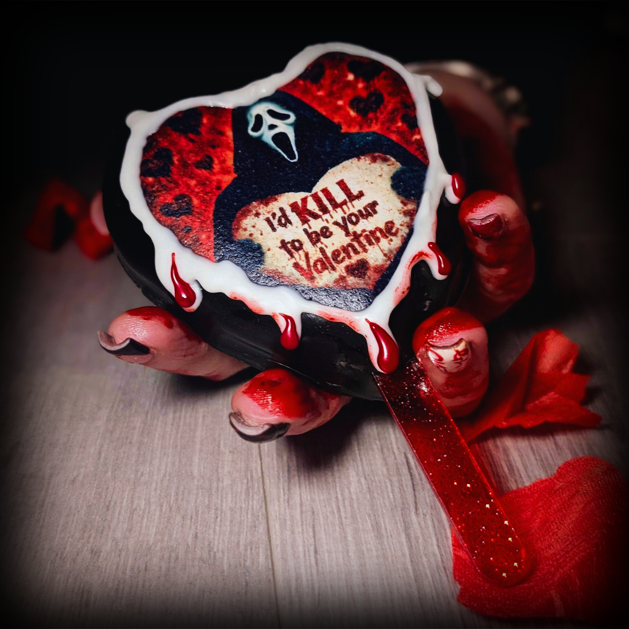 A black heart shaped cake inspired by ghostface from scream. the heart has a white and red chocolate drip which is being held by a bloodied hand