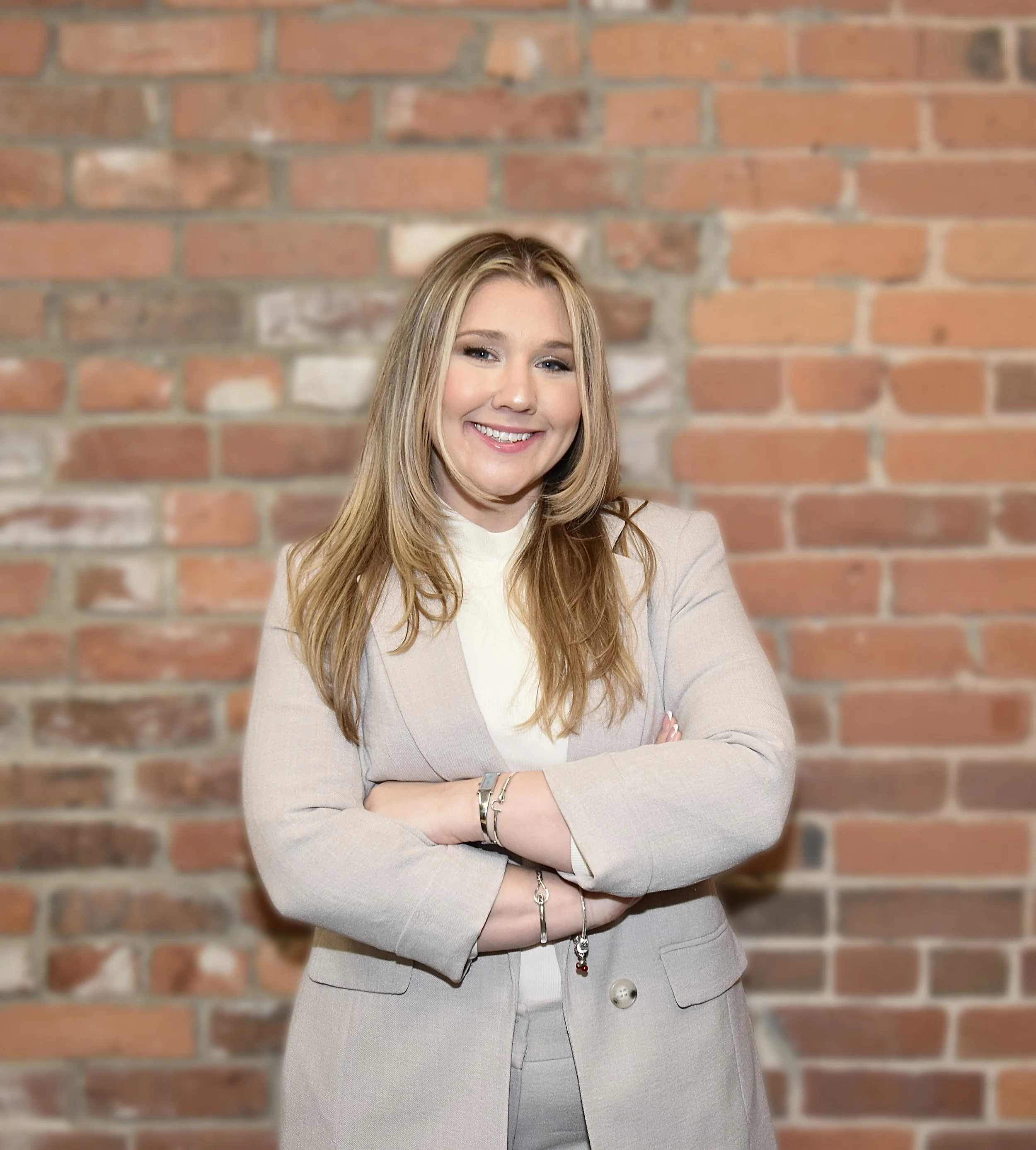 A woman with long blonde hair wearing a beige blazer and gray pants, smiling with crossed arms, standing in front of a brick wall.