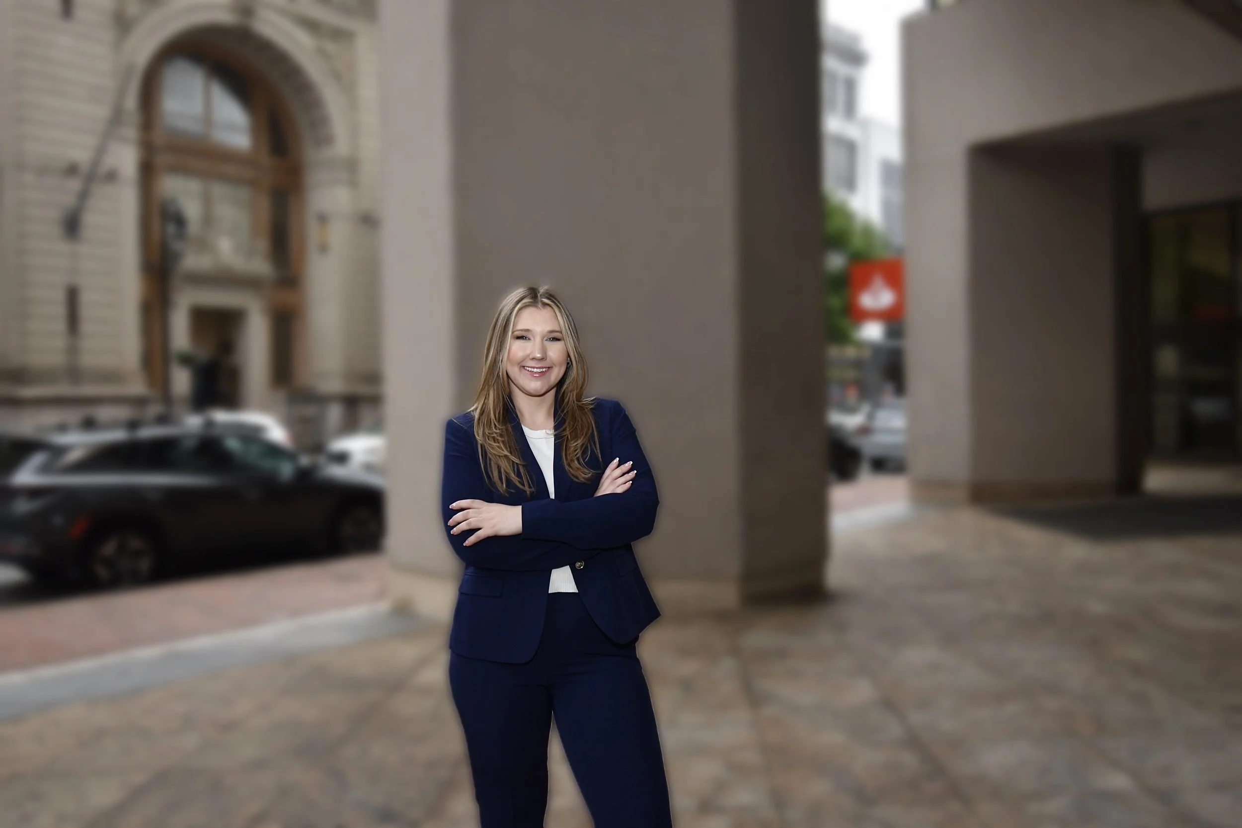 A smiling woman with long blonde hair, wearing a dark blazer and white shirt, standing outdoors with arms crossed in front of a beige building and parked cars.