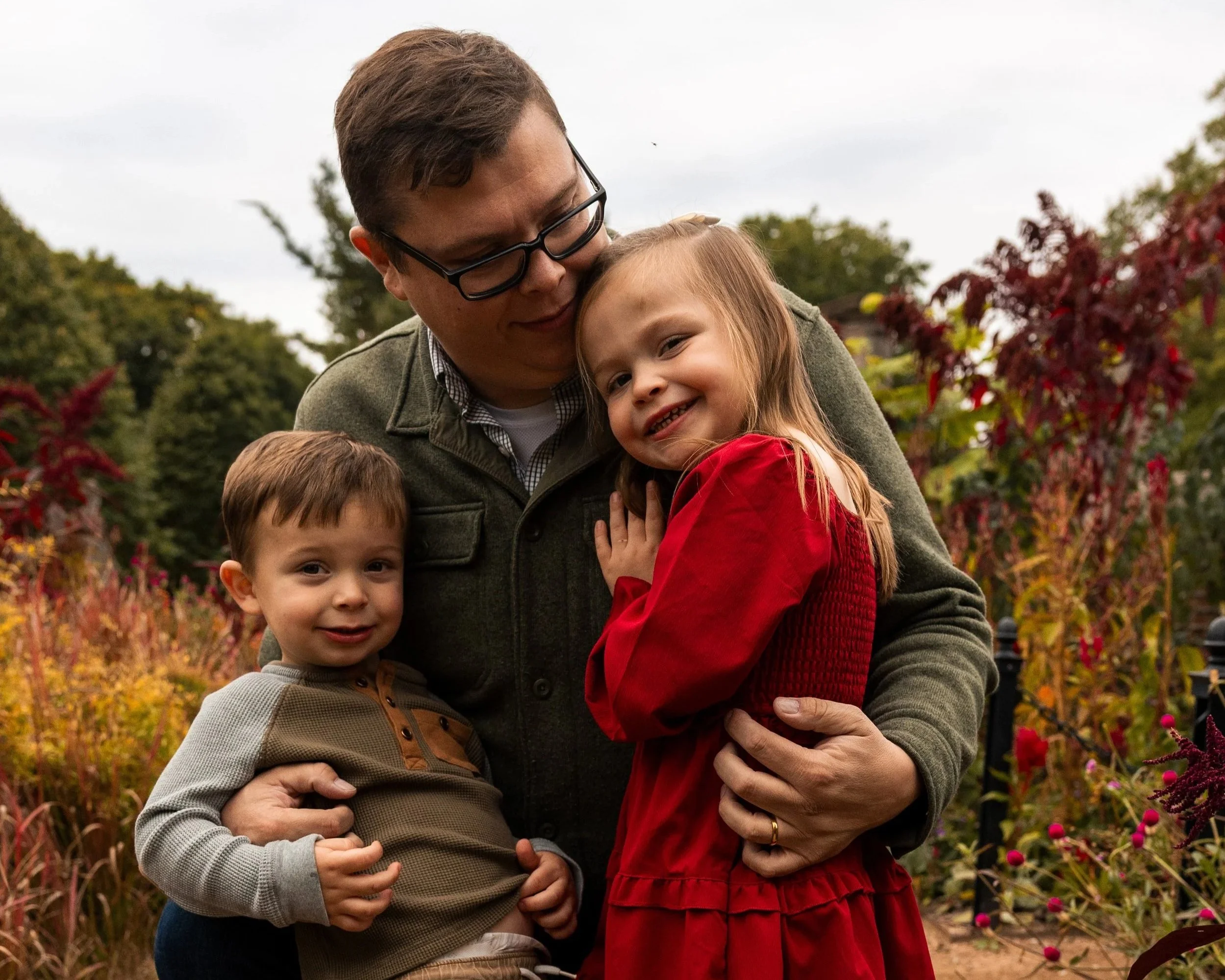 A man with glasses, holding two children, stands outdoors in a garden with colorful plants and trees. The man has dark hair, and the children are smiling, with the girl wearing a red dress and the boy in a beige and gray shirt.