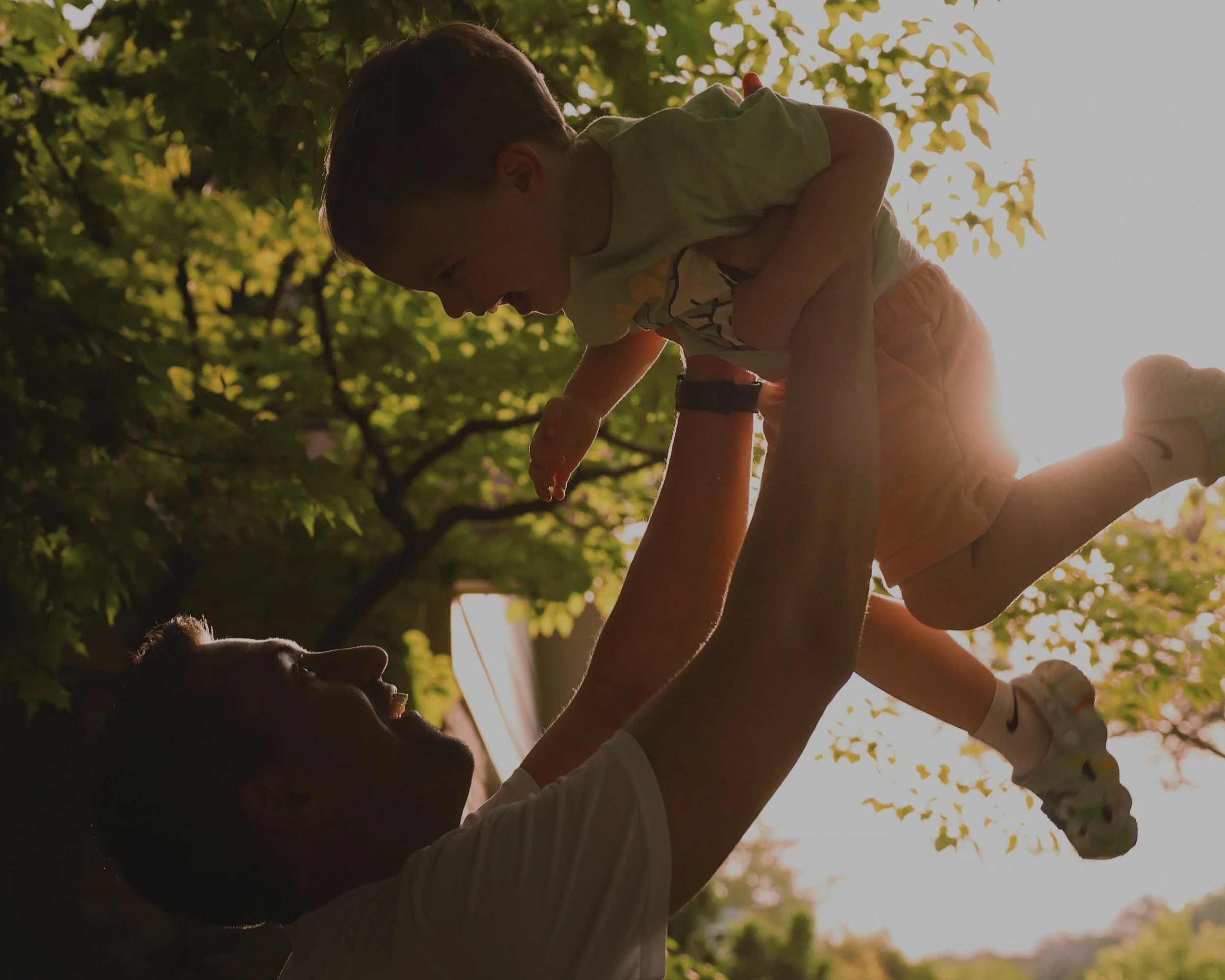 A person lifting a young child into the air outdoors with trees and sunlight in the background.