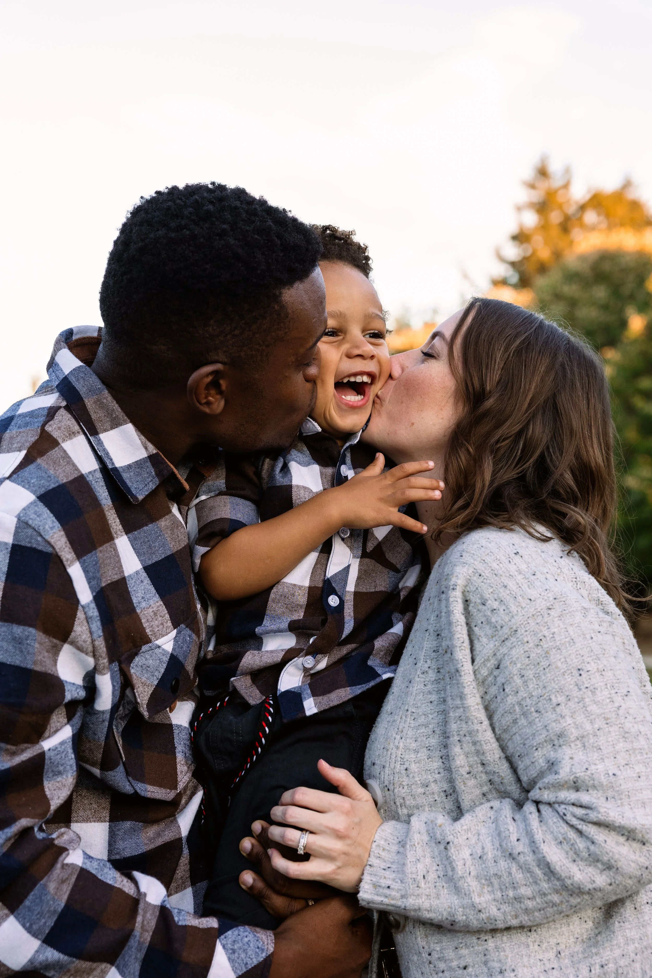 A family of three, a man, woman, and young boy, outdoors during sunset. The woman is kissing the boy on the cheek, who is smiling and laughing. The man is holding the boy, and the woman has her hand on his waist. All are wearing plaid shirts and appear happy.