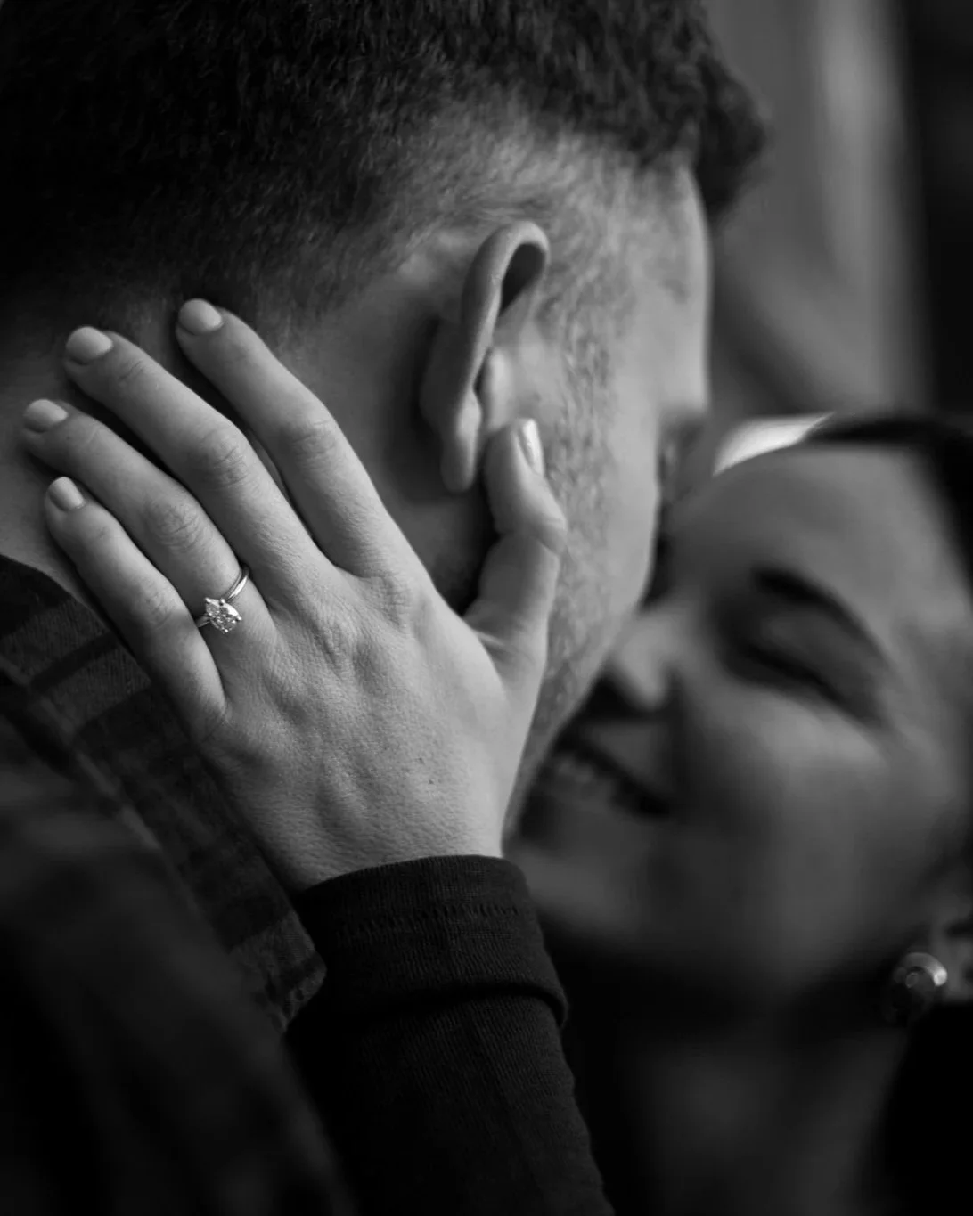 A close-up black and white photo of a person touching to their ear, with a focus on their hand and wedding ring. The person has short hair and is resting their head on their hand, slightly tilted.