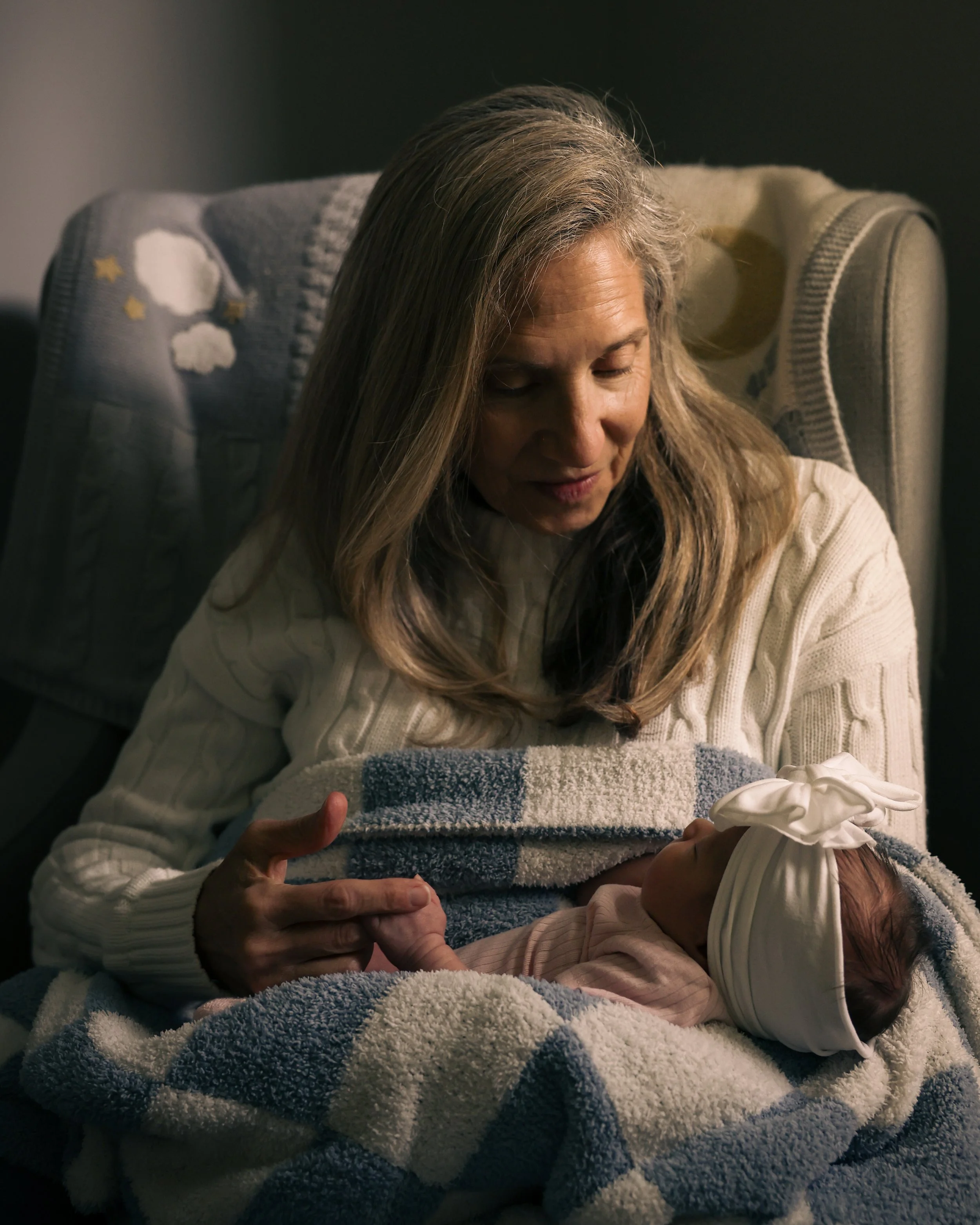 A woman with gray hair looking at a newborn baby wrapped in a towel, wearing a headband. The woman is sitting in a chair with a blanket behind her.