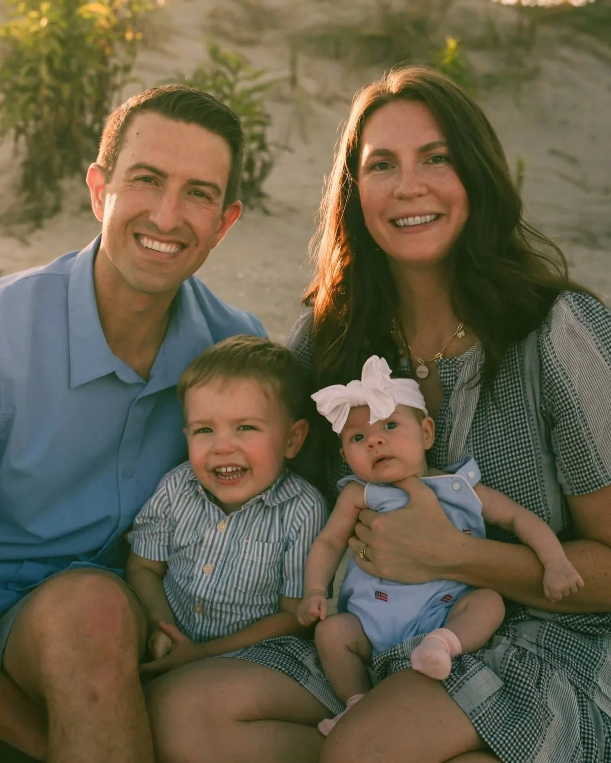 A family of two adults and two children smiling at the camera outdoors during sunset, sitting on a sandy area with sparse greenery in the background.
