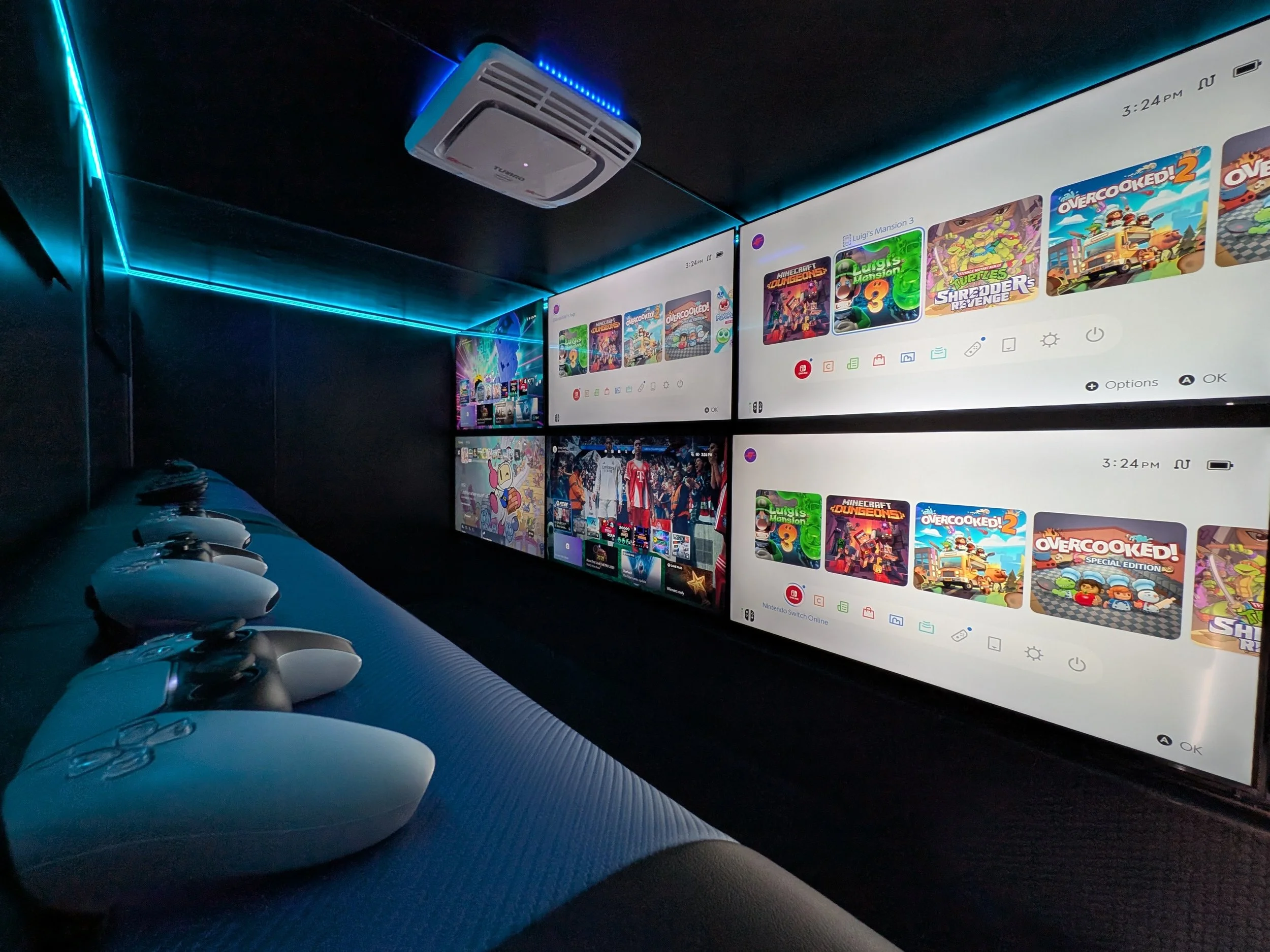 A gaming setup inside of a Jersey Gamer mobile video game truck with two large monitors displaying a Nintendo Switch home menu with various game icons. Below the monitors, there are several white Nintendo Switch controllers lined up on a dark, textur