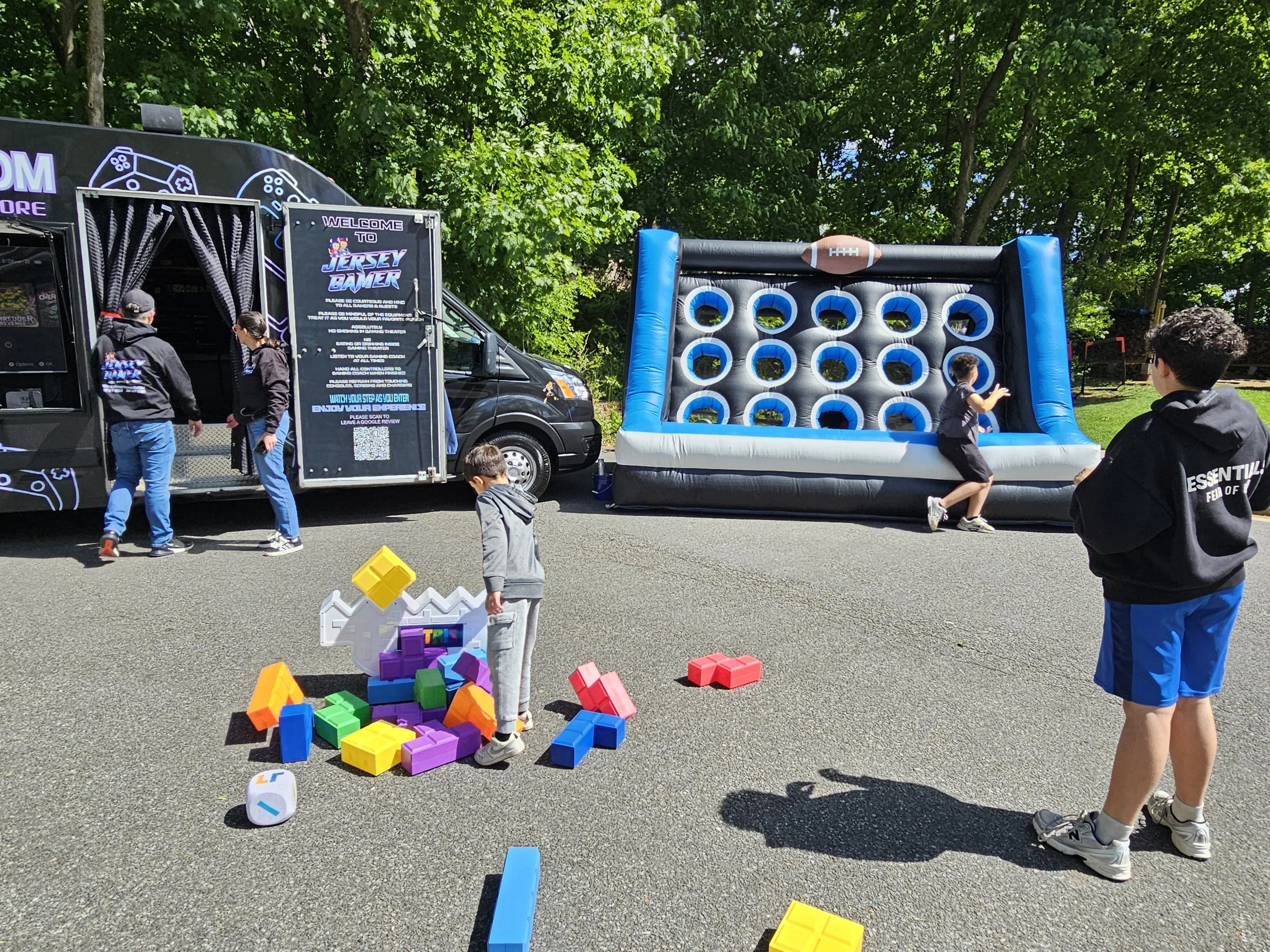 Several people enjoying a variety of outdoor games, some inflatable, during a party outside of a Jersey Gamer mobile video gaming truck,.