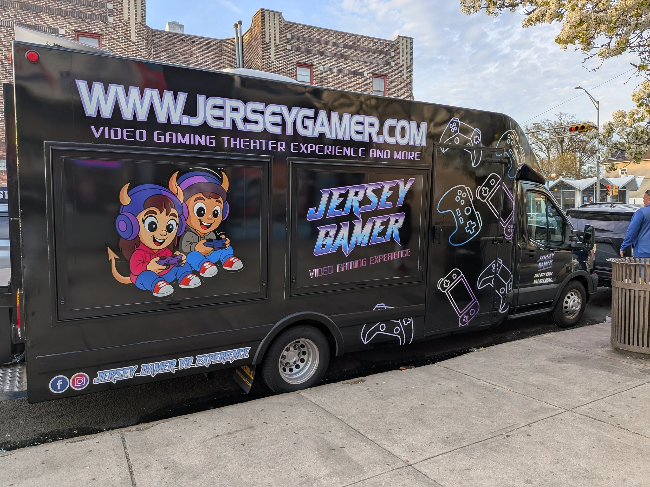 Black mobile advertising truck with colorful graphics, featuring cartoon characters of two kids wearing headphones playing video games, and text promoting JerseyGamer.com for video gaming experiences, with illustrations of game controllers and social