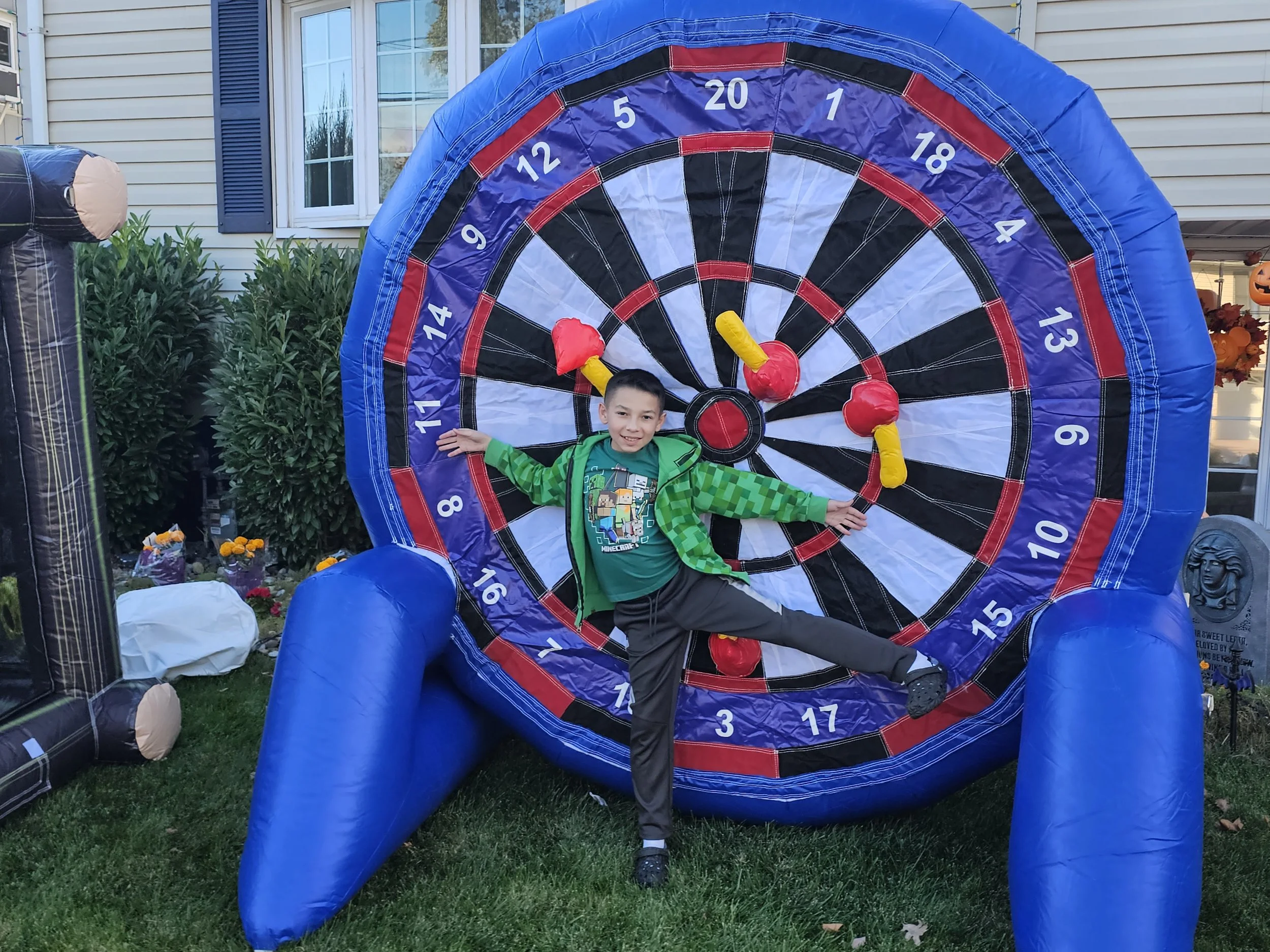 A young boy is standing in front of an inflatable dartboard, holding a toy axe in each hand. The scene appears to be outdoors in a yard, with a house and greenery in the background. The boy is smiling and wearing a green jacket and a Minecraft t-shirt.
