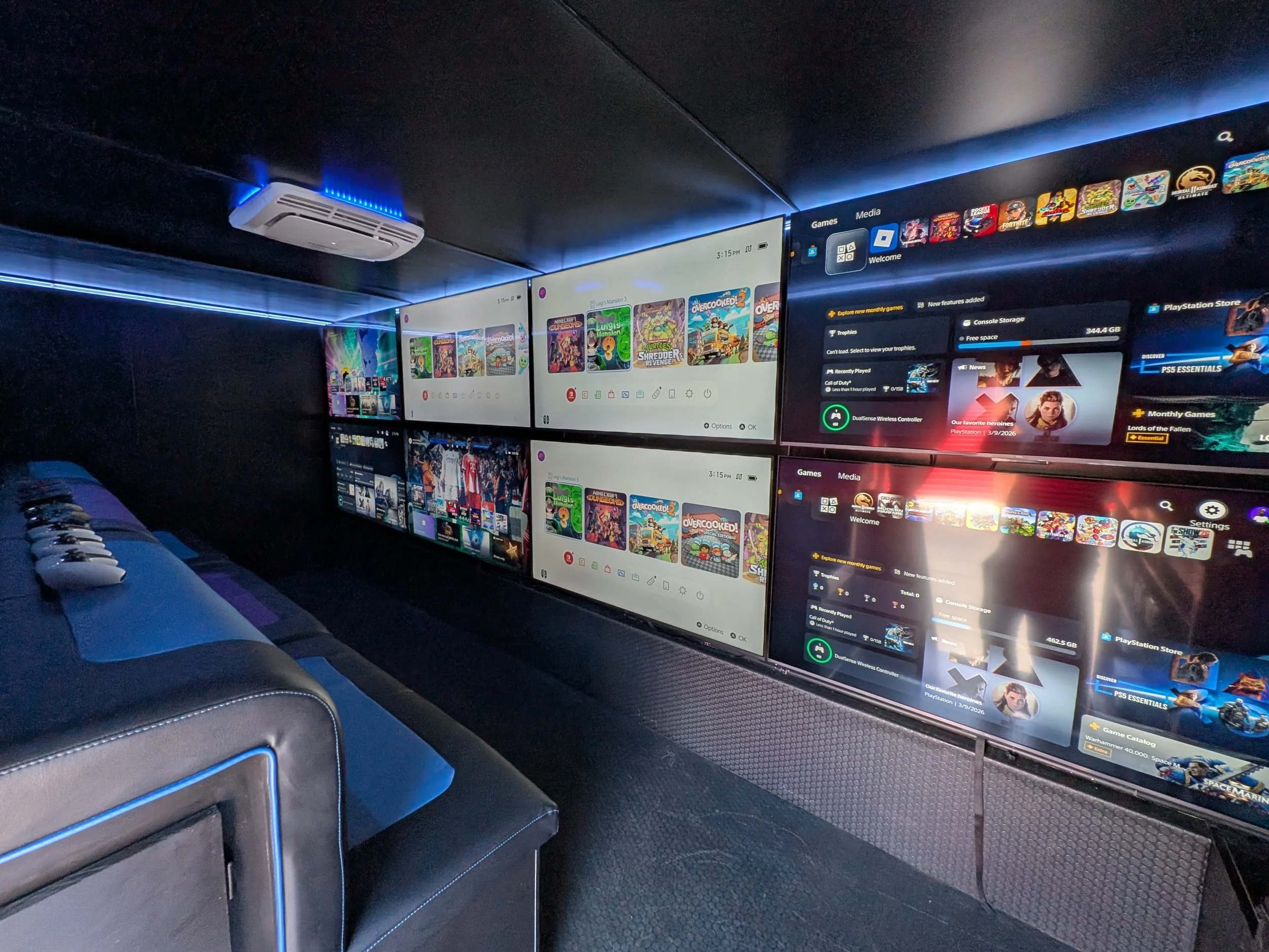 Video game truck interior with multiple monitors displaying a gaming menu and game library, blue LED lighting, and a black ceiling with an air conditioning unit.