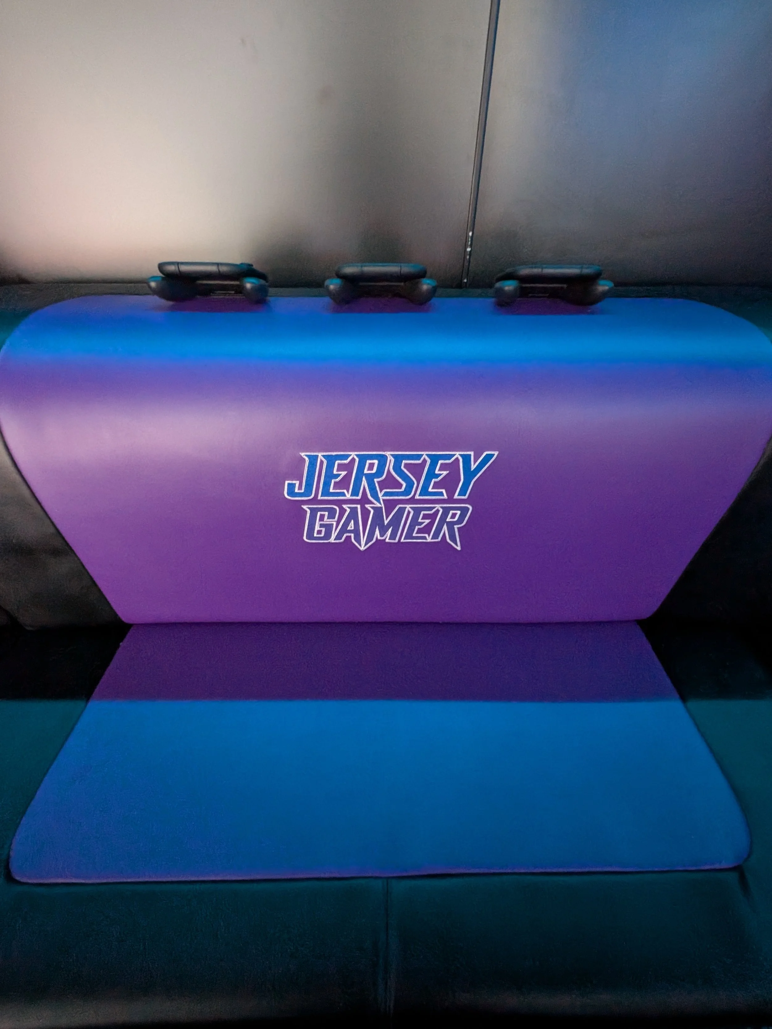 Purple and blue seat inside a video game truck with 'Jersey Gamer' logo on front, three remote controls placed on top.