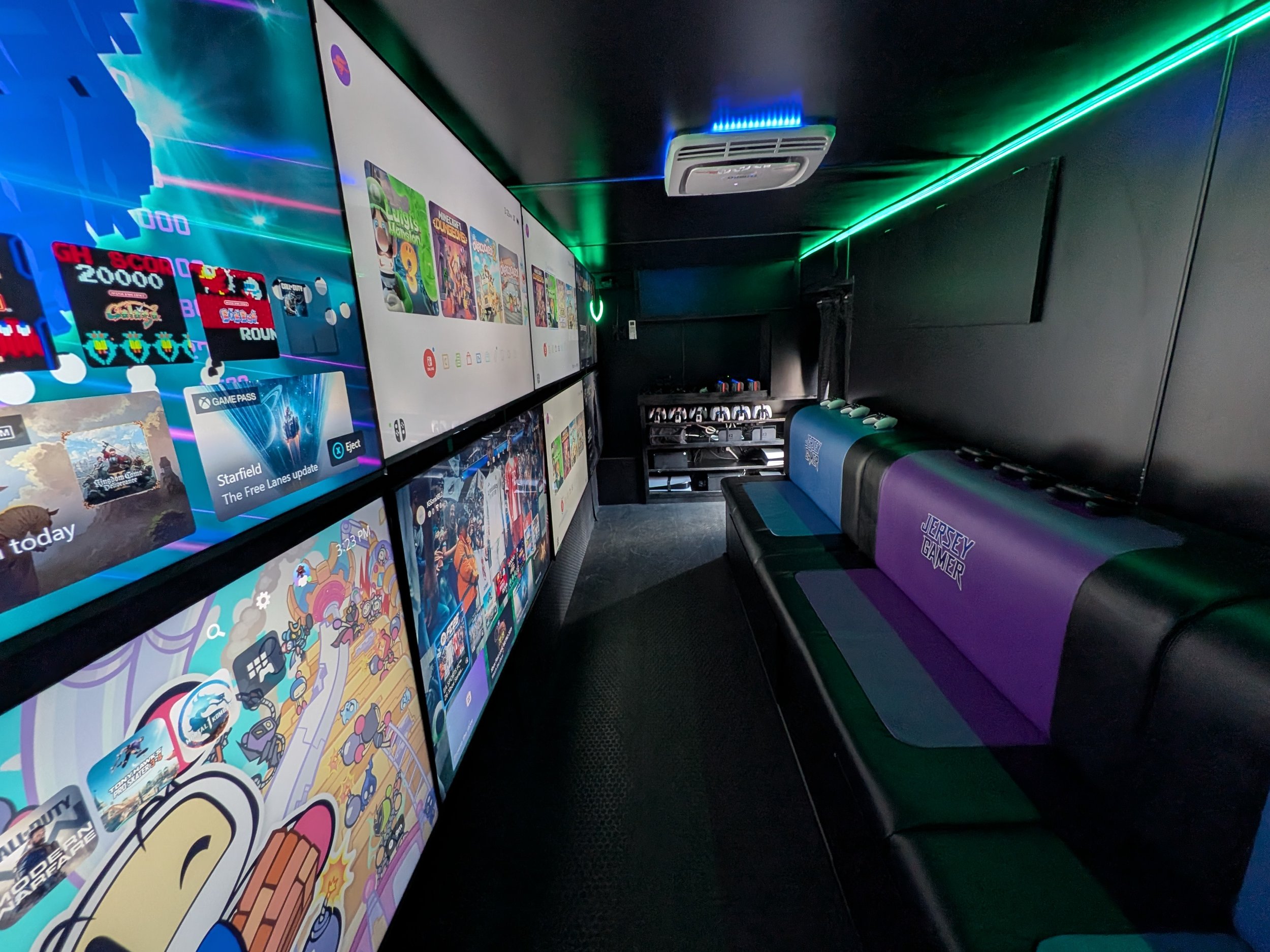 Video game truck interior with a row of large screens displaying video games or apps, purple and green neon lighting, black walls, and a bench with purple and teal seating labeled Jersey Gamer.