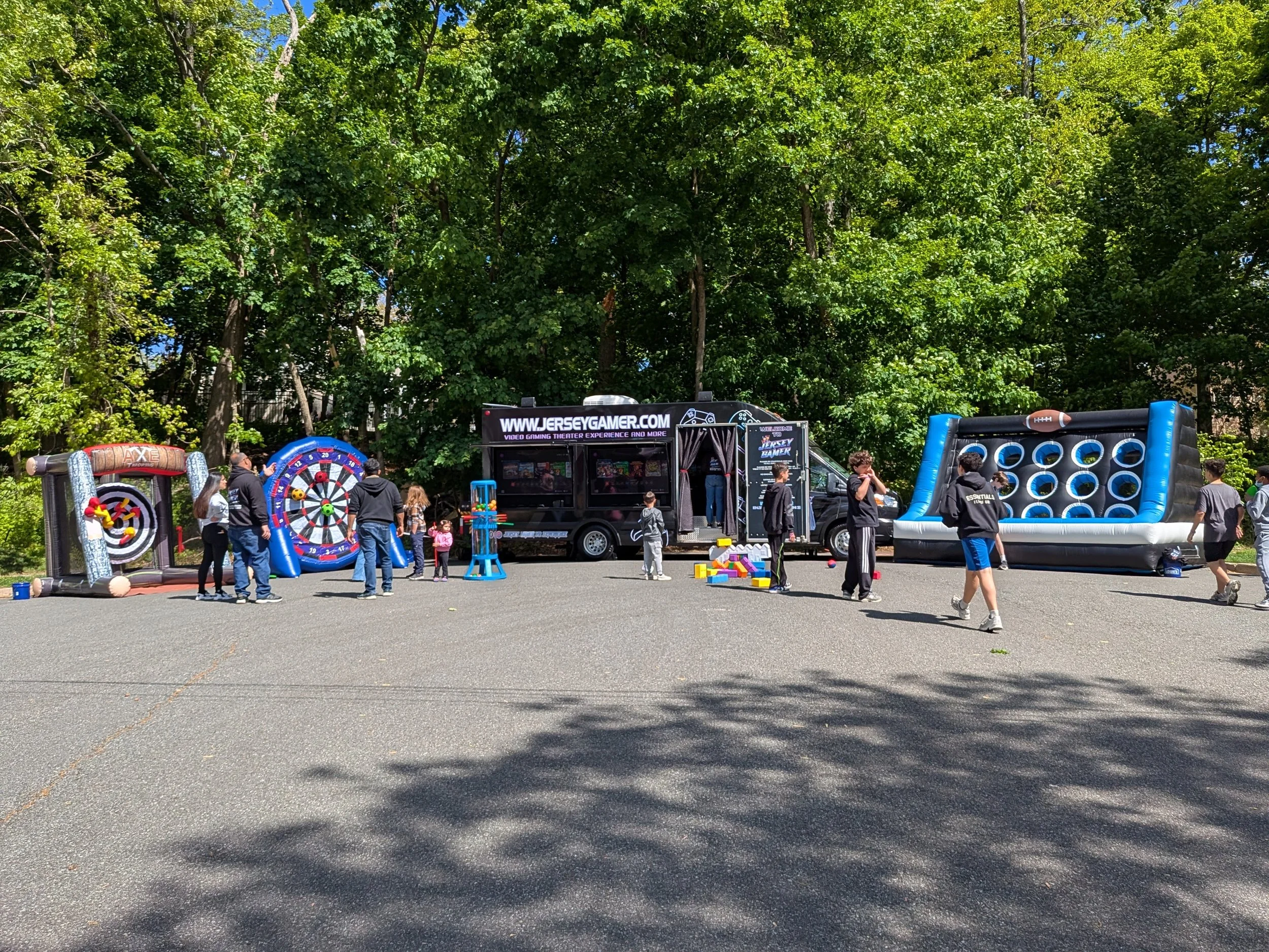 People enjoying a fully set up Jersey Gamer outdoor party.  The party includes a Jersey Gamer mobile video game truck, stationary outdoor games and three inflatable outdoor games - soft axe throwing, soccer darts, and football toss.