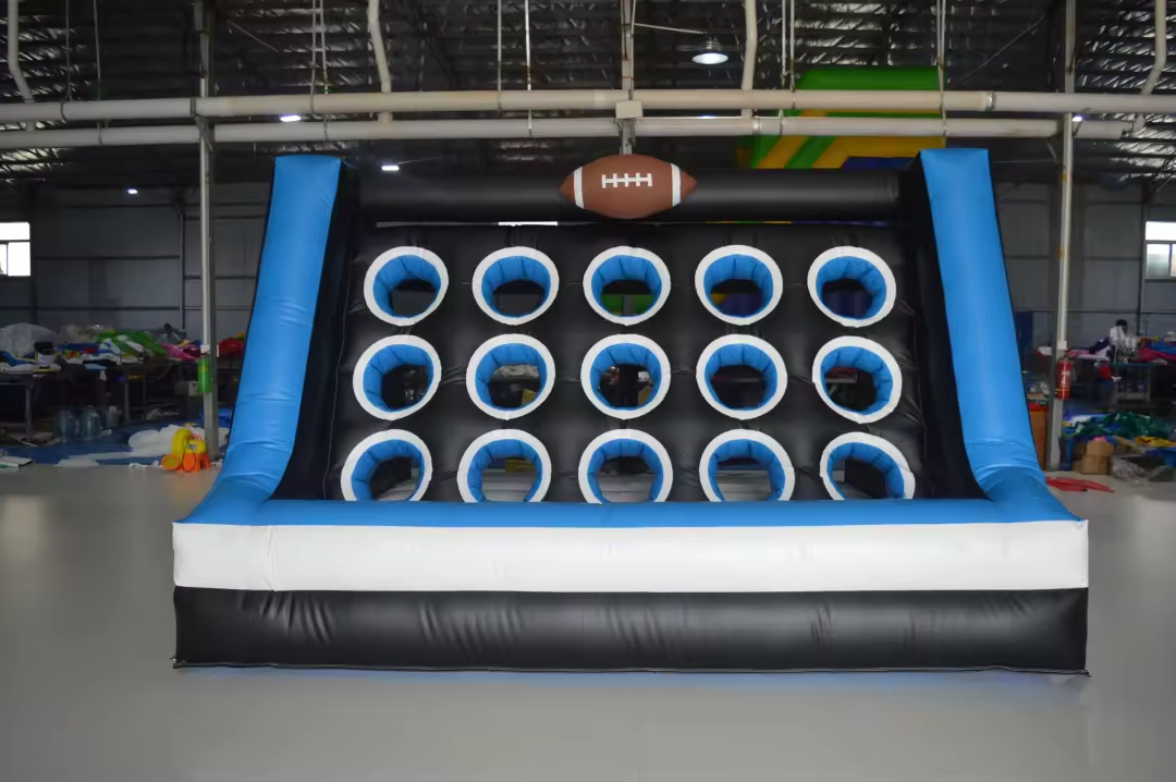 An inflatable game featuring a football theme, with a black, blue, and white color scheme, multiple circular openings, and a brown inflatable football on top, situated indoors with other inflatables and equipment in the background.