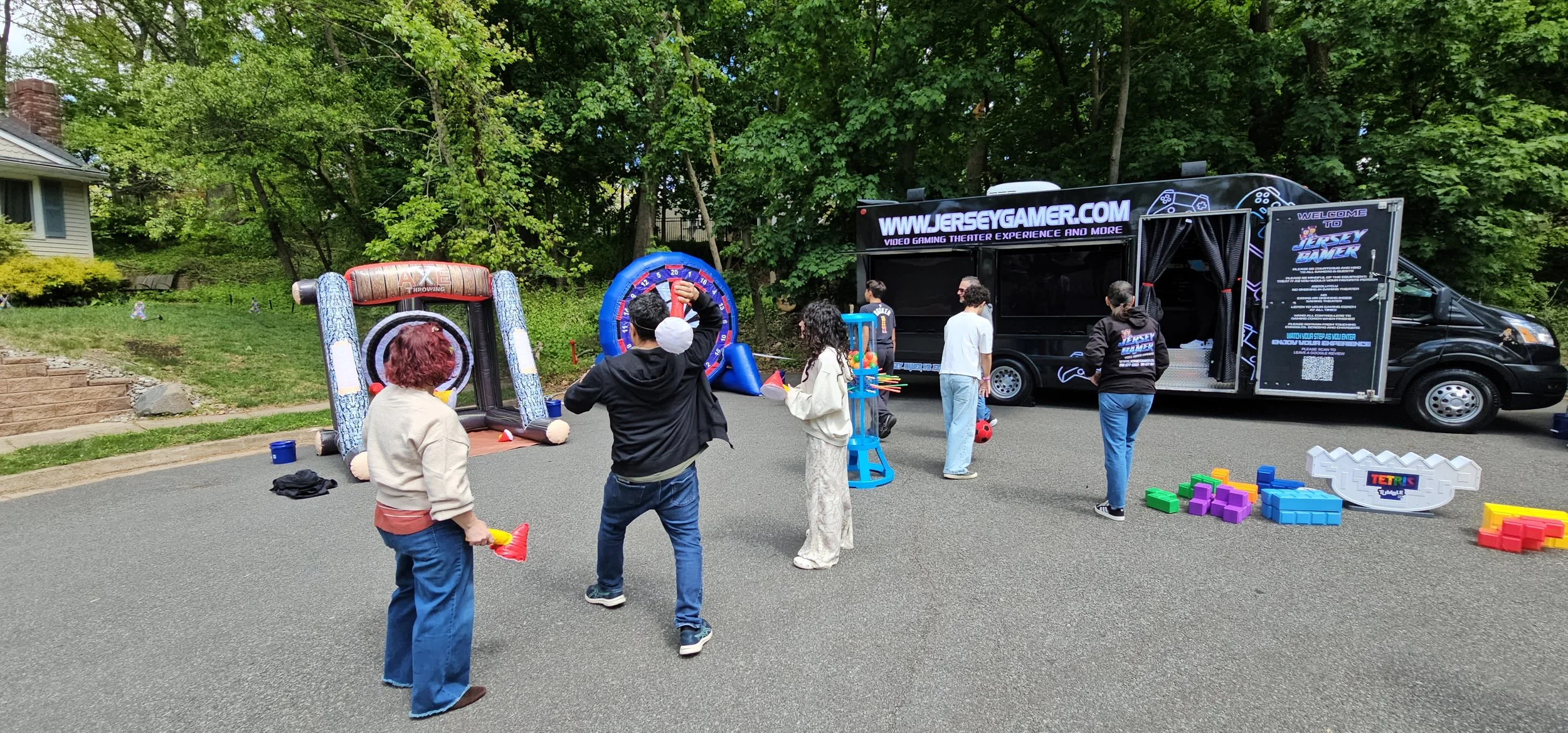 Several people enjoying inflatable outdoor games, one of them soft axe throwing and another called soccer darts, outside of a Jersey Gamer mobile video game truck.