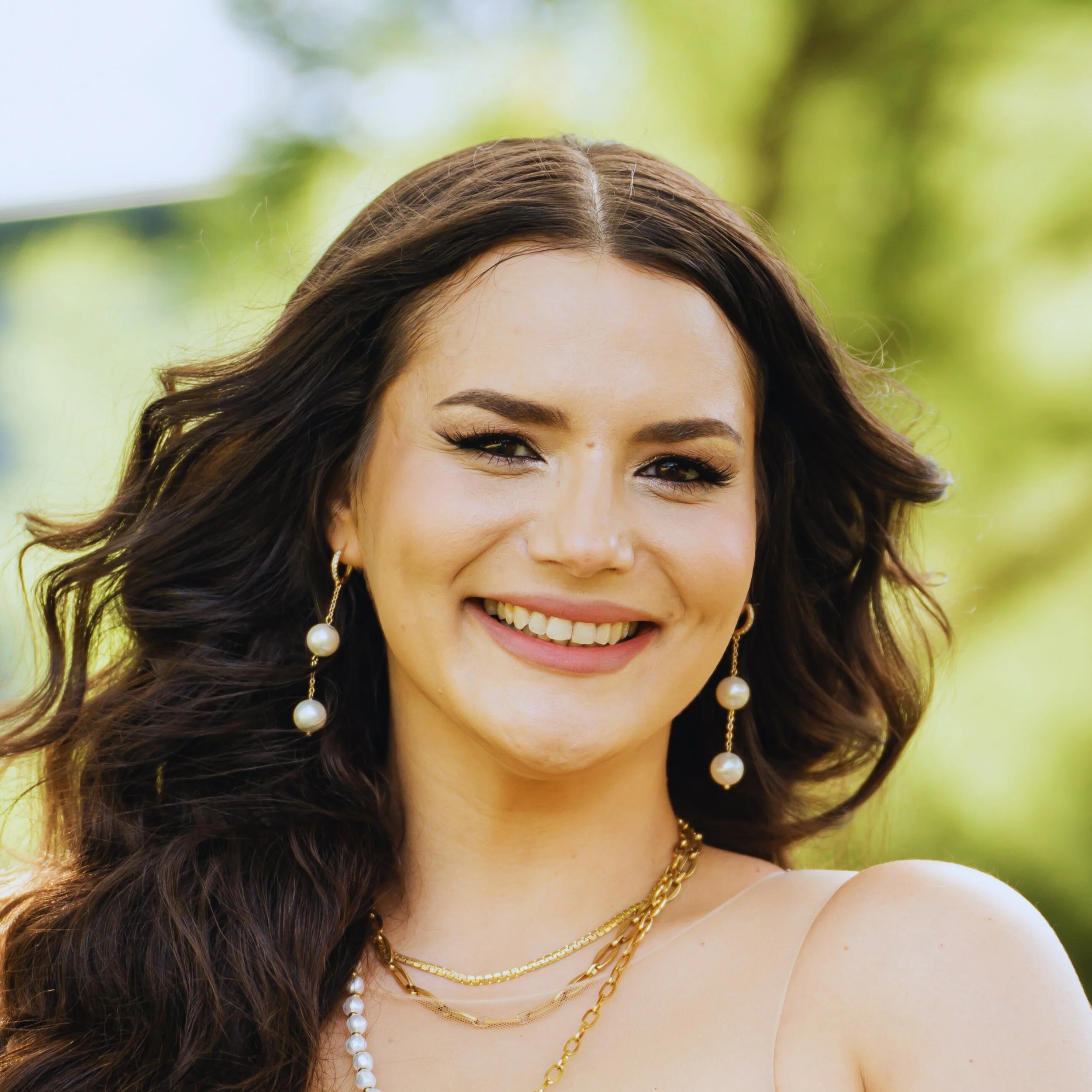 Close-up of a woman with long wavy brown hair, wearing pearl drop earrings, layered gold necklaces, and a beige top, smiling outdoors with a blurred green background.