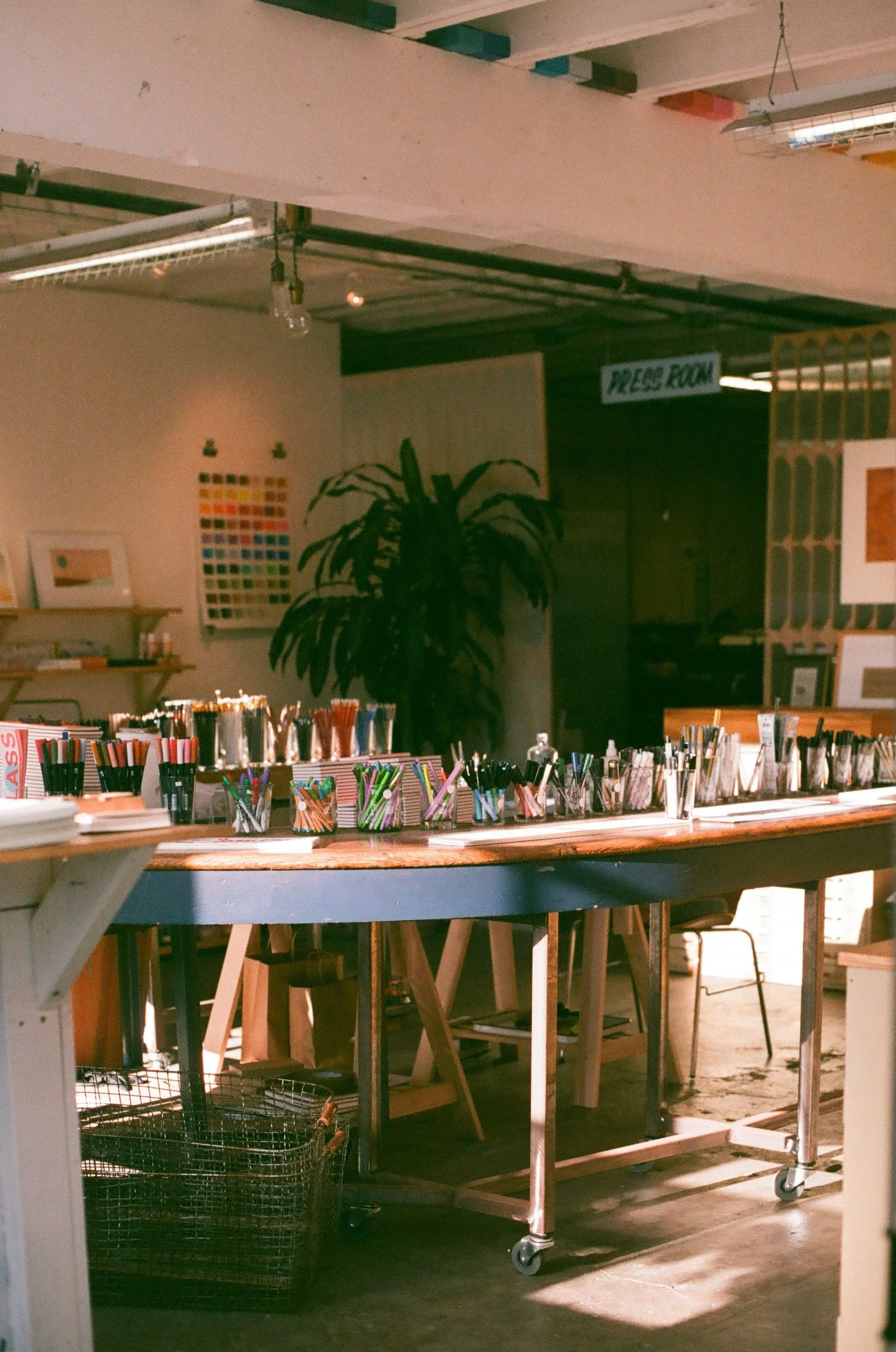 An indoor art studio with a wooden table filled with various colored pens, markers, and art supplies. There is a large potted plant in the background and artwork on the walls. A sign reads 'PRESS ROOM' in the background.