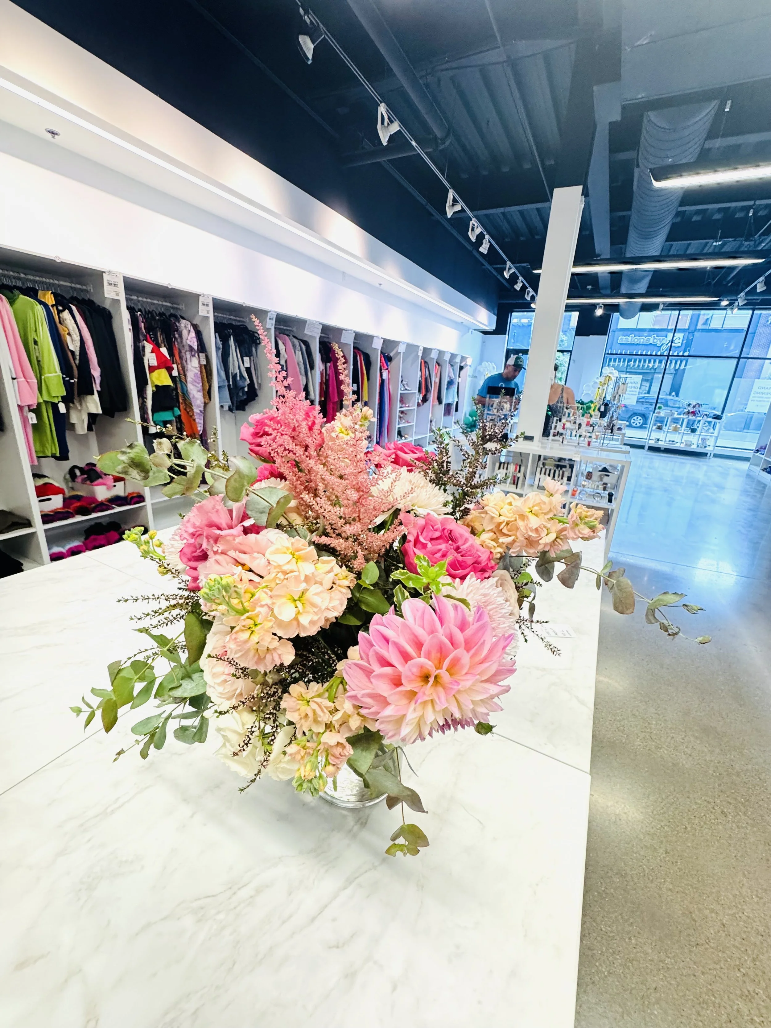 Pink and peach floral arrangement on a white table inside a retail store with clothing on racks and large windows showing outside view.