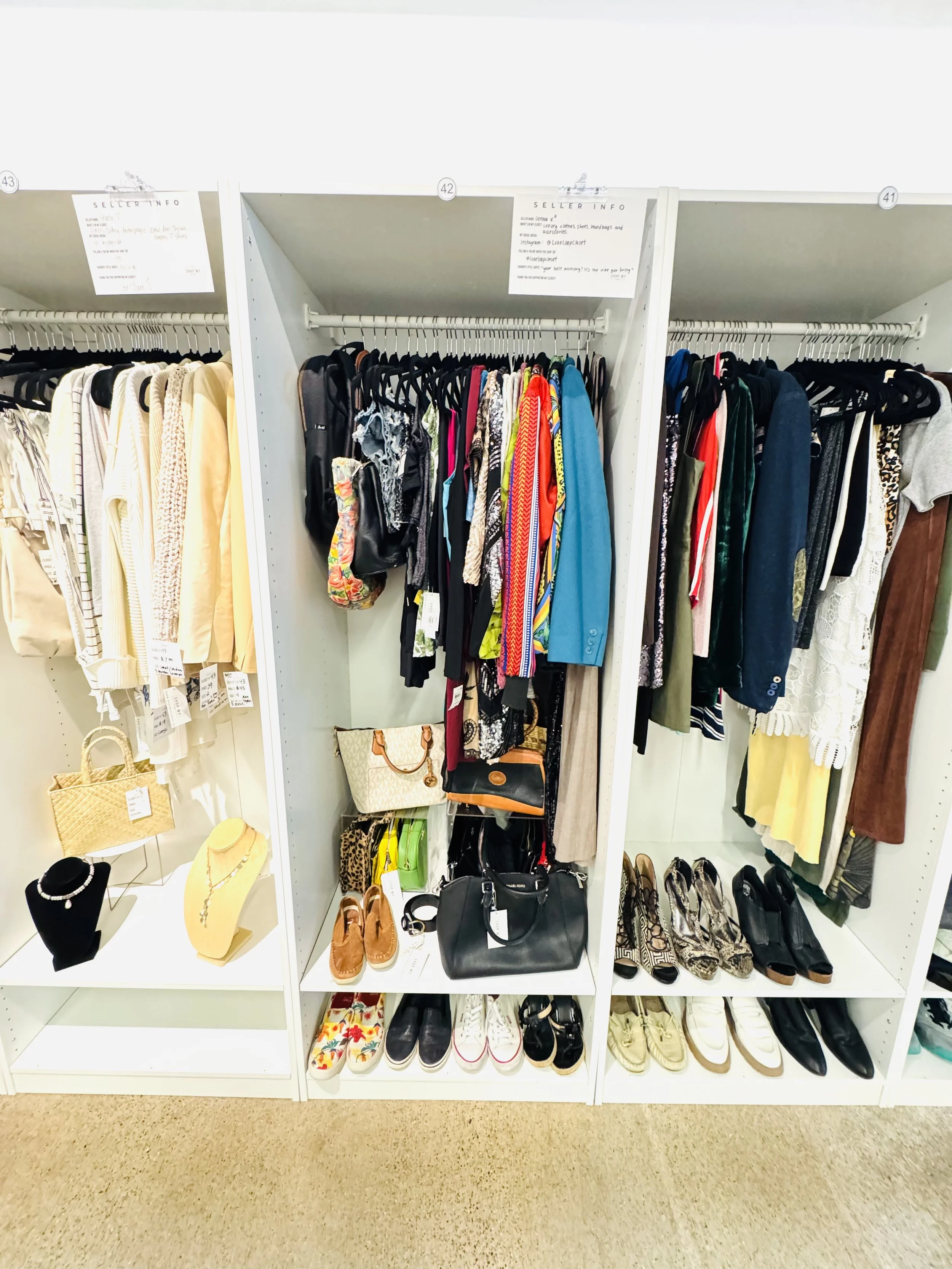 Clothing display in a thrift store with various dresses, skirts, shoes, purses, and jewelry on white shelves and racks.