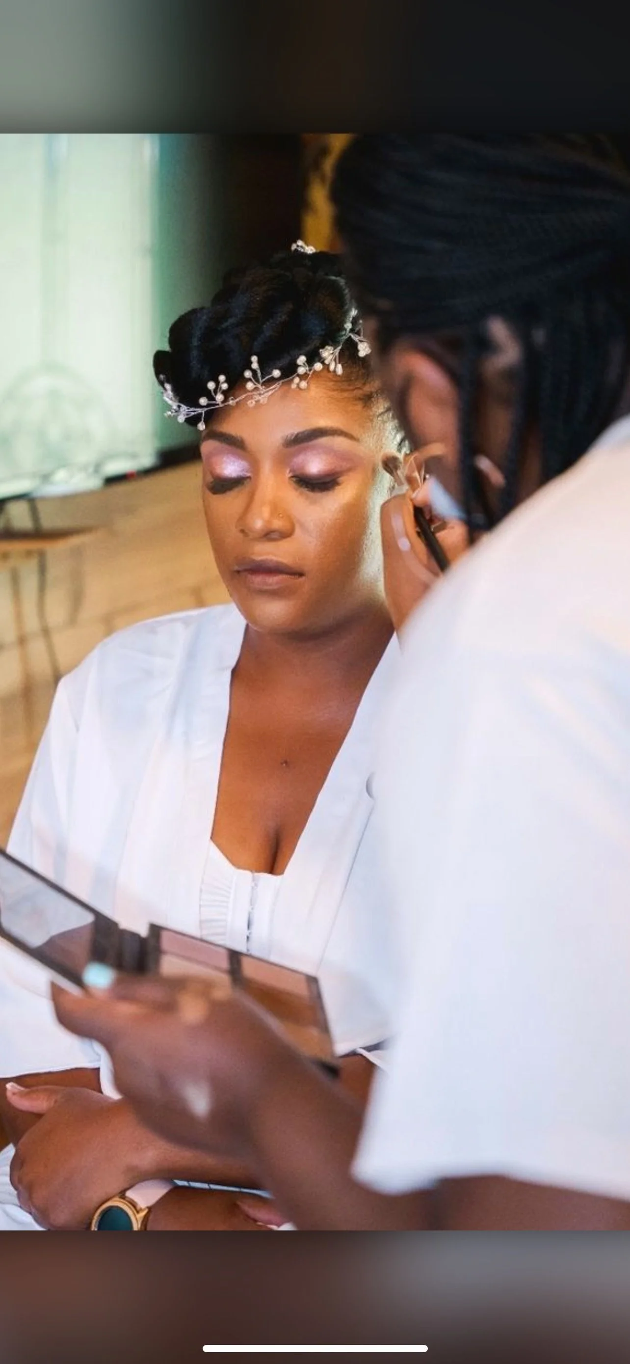 A woman with a decorative headpiece and pink eyeshadow gets her makeup done by a makeup artist.