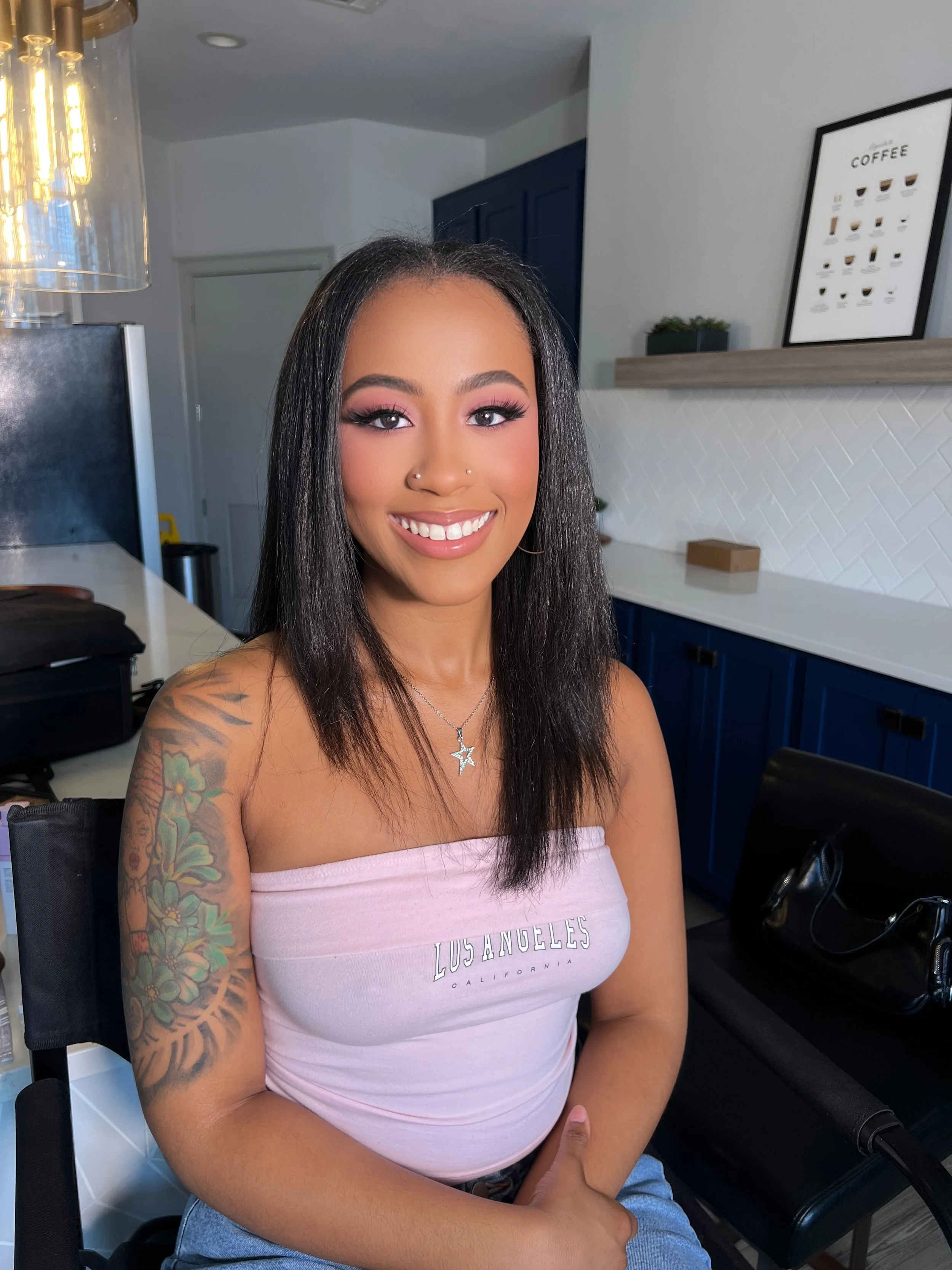 A young woman with long black hair and tattoos on her left arm, sitting in a chair with a smile, wearing a pink tube top with the text 'Los Angeles California,' and silver star-shaped necklace, in a modern kitchen with blue cabinets and a framed coffee poster on the wall.
