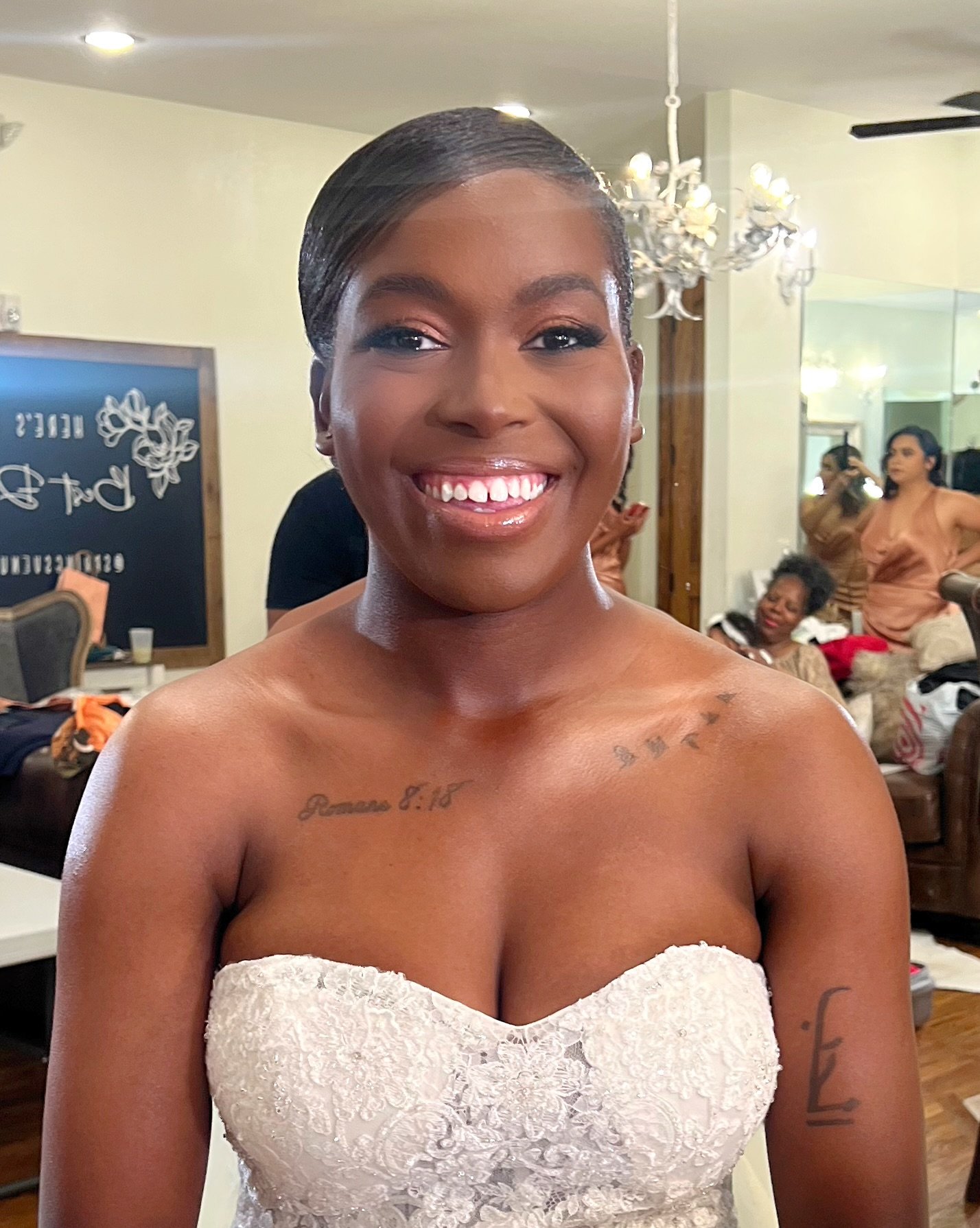 Smiling woman with short, slicked-back hair taking a selfie in a dressing room or backstage area, with other women and a chandelier visible in the background.