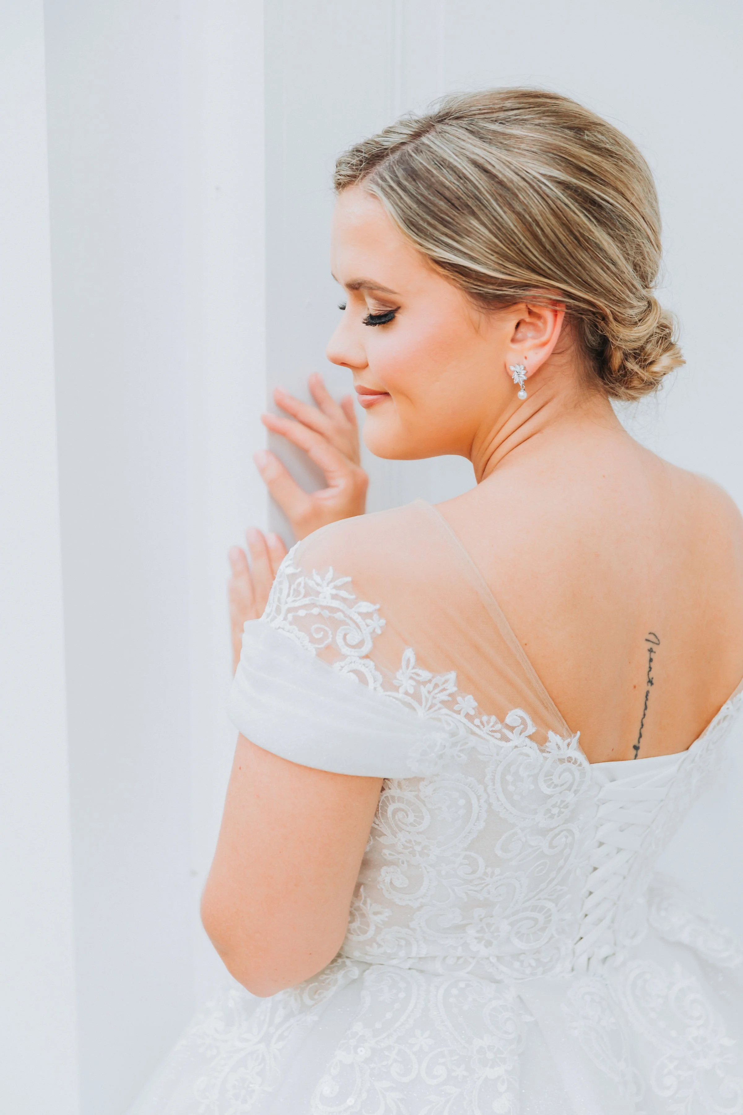 Side profile of a bride in a white lace wedding dress, with her eyes closed, smiling softly, touching a white wall, wearing floral earrings and a low bun hairstyle.