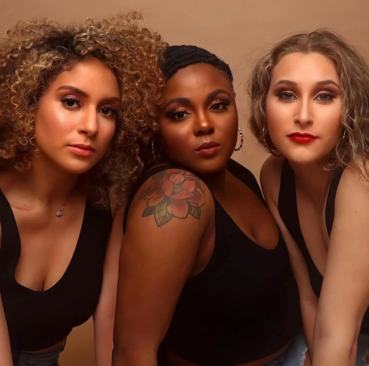 Three women with different hairstyles and makeup posing closely together, wearing black tops and jewelry, against a plain background.