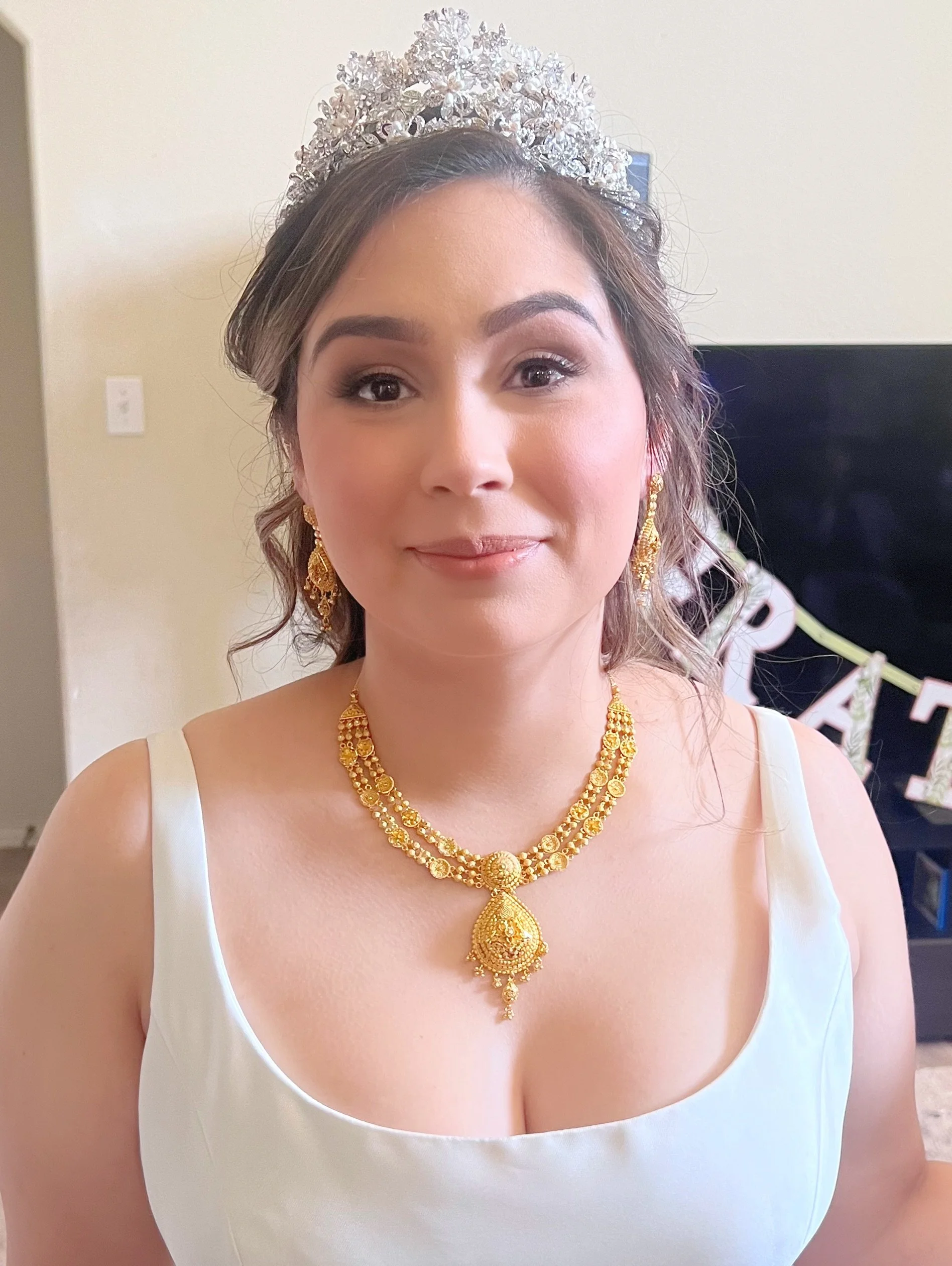 A woman wearing a tiara and gold jewelry, dressed in a white dress, taking a selfie indoors.