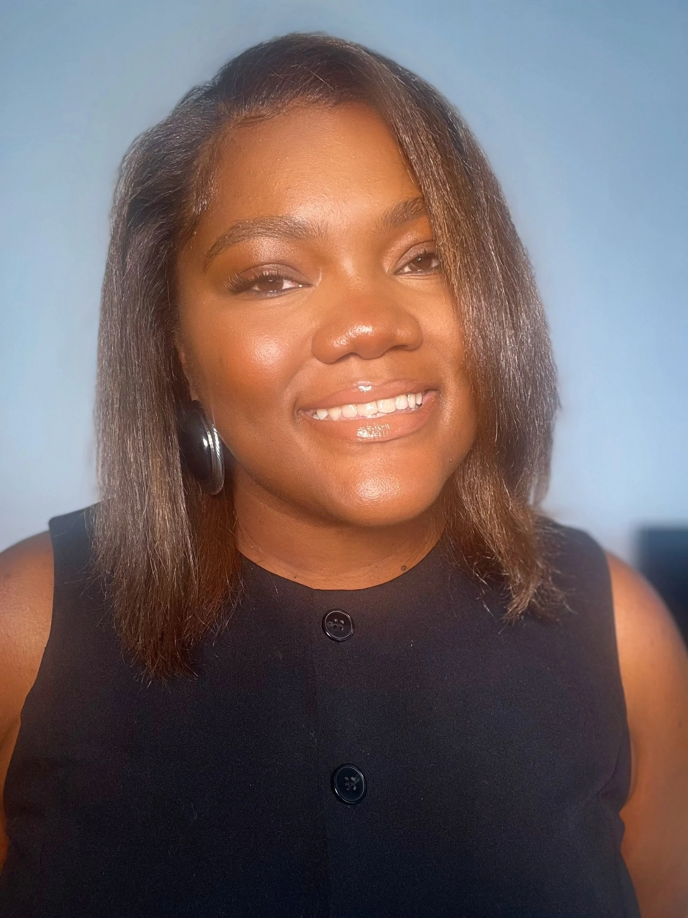 Close-up of an African American woman smiling, wearing a sleeveless black top with black buttons, and large hoop earrings, against a light blue background.