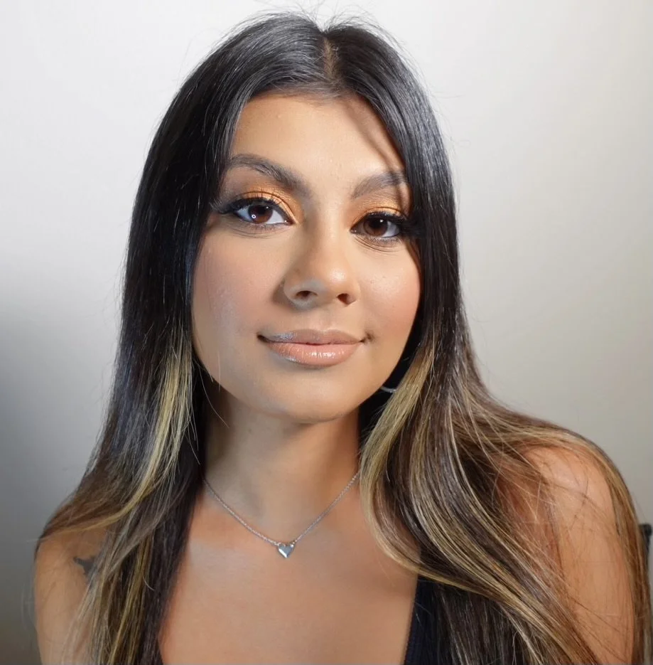 Close-up portrait of a woman with long, dark hair and light brown highlights, wearing a necklace with a heart pendant, makeup with orange eyeshadow and glossy lips, smiling slightly, against a plain light background.
