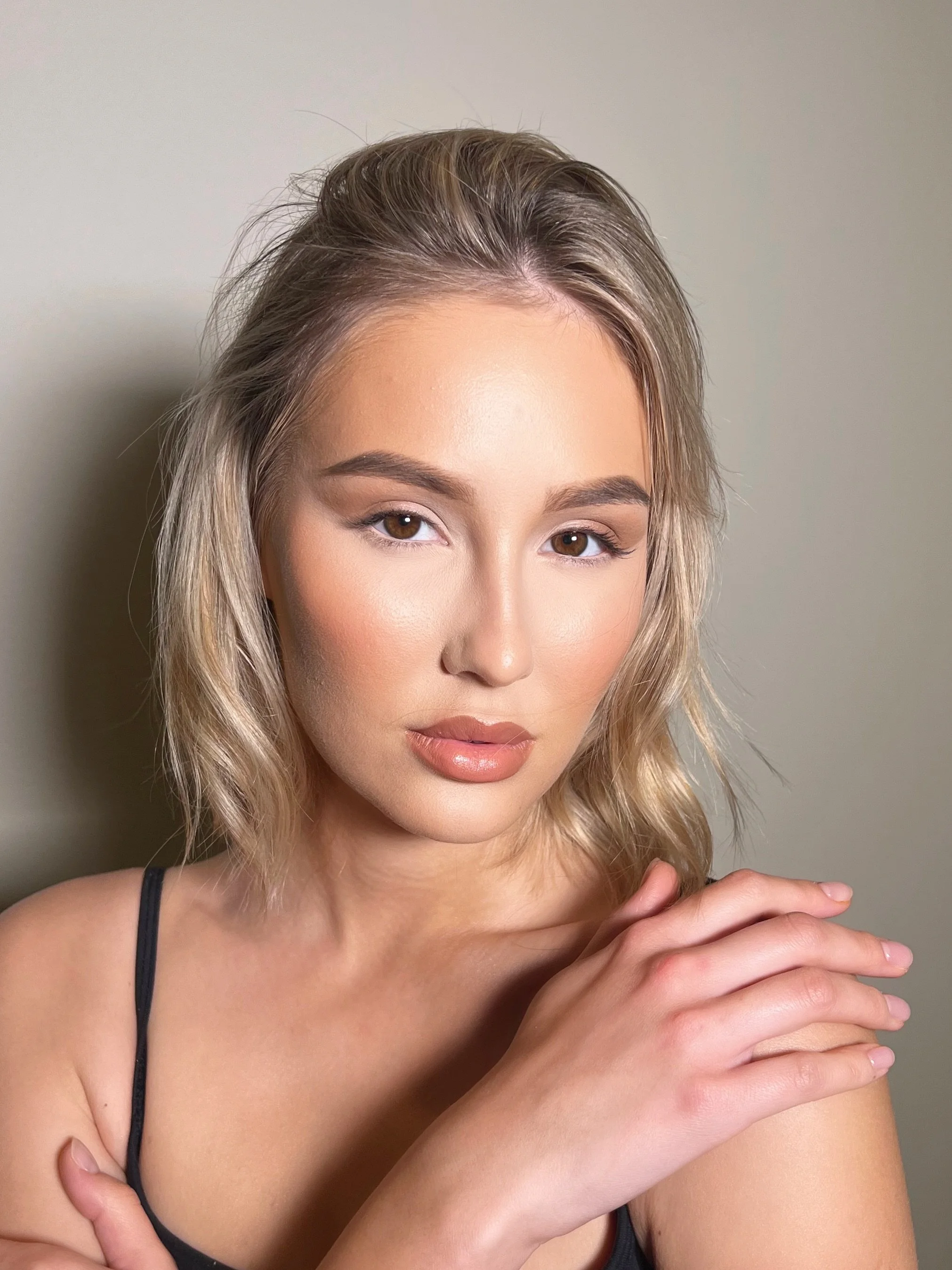 Close-up of a woman with blonde hair and brown eyes, wearing a black strap camisole, with her hand resting on her shoulder against a plain light gray background.