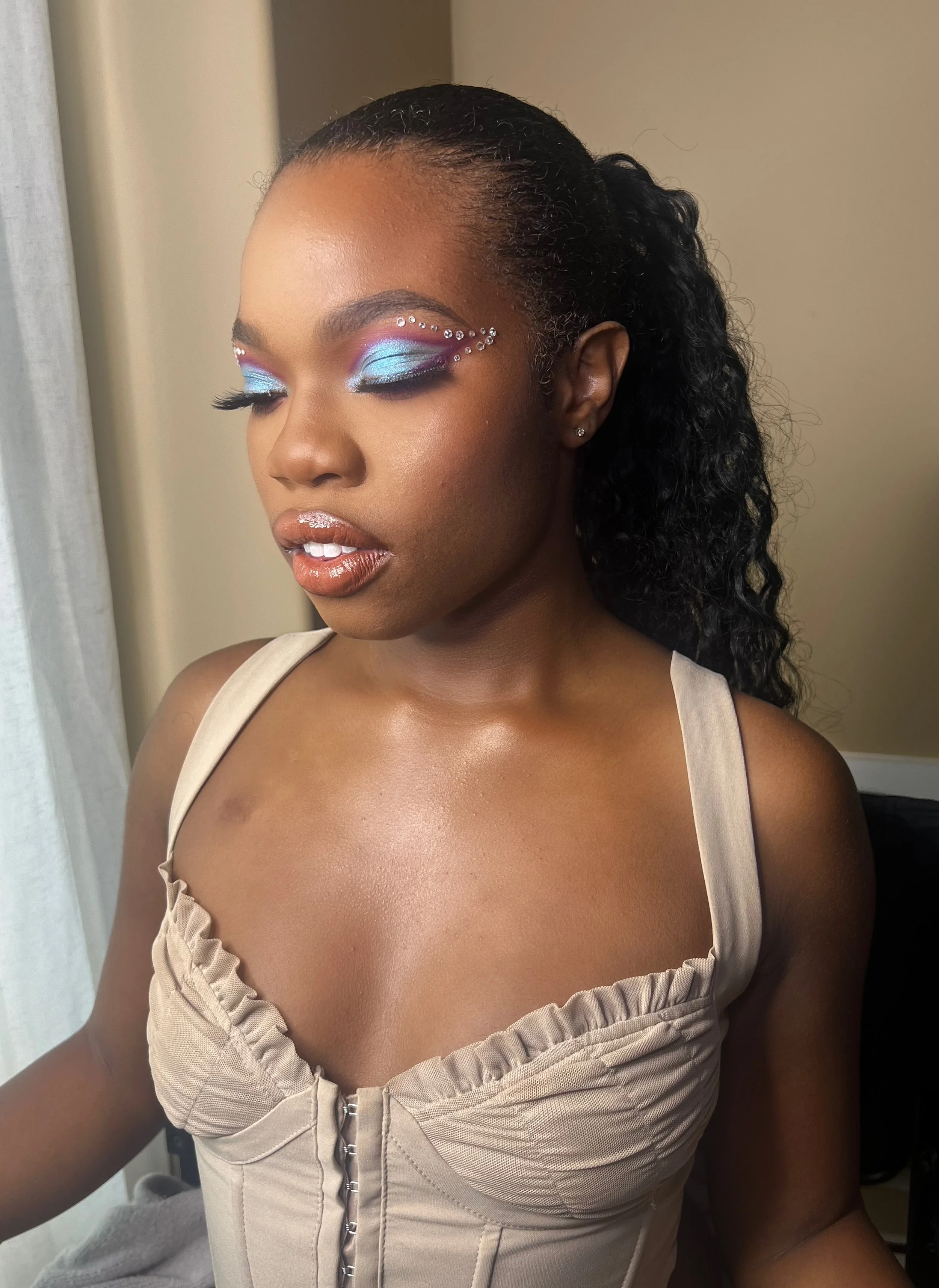 Close-up of a woman with artistic makeup, including blue and purple eyeshadow, rhinestones around her eyes, and glossy lips, wearing a beige dress with ruffle details.