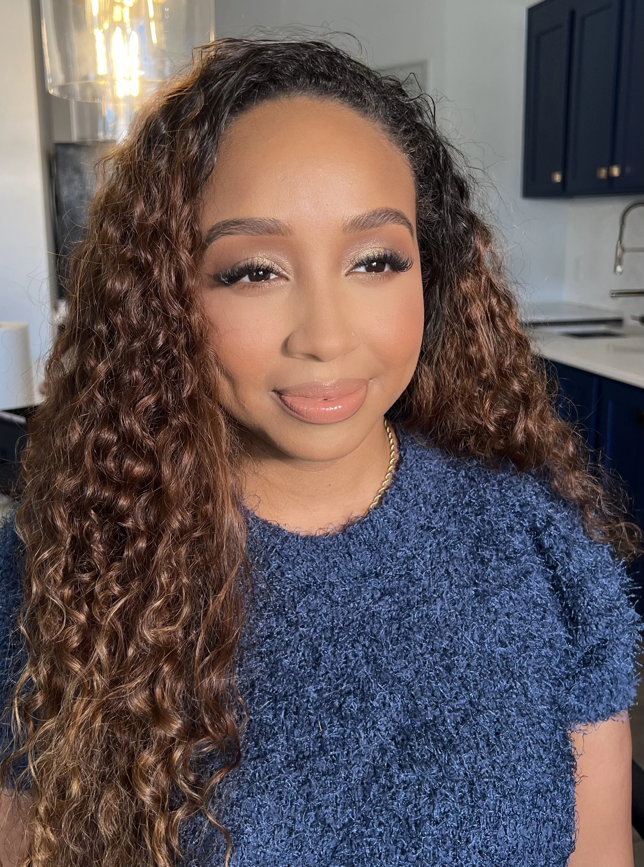 Close-up of a woman with long, curly hair and makeup, wearing a blue sweater and a gold chain necklace, indoor setting with kitchen cabinets and sink in the background.