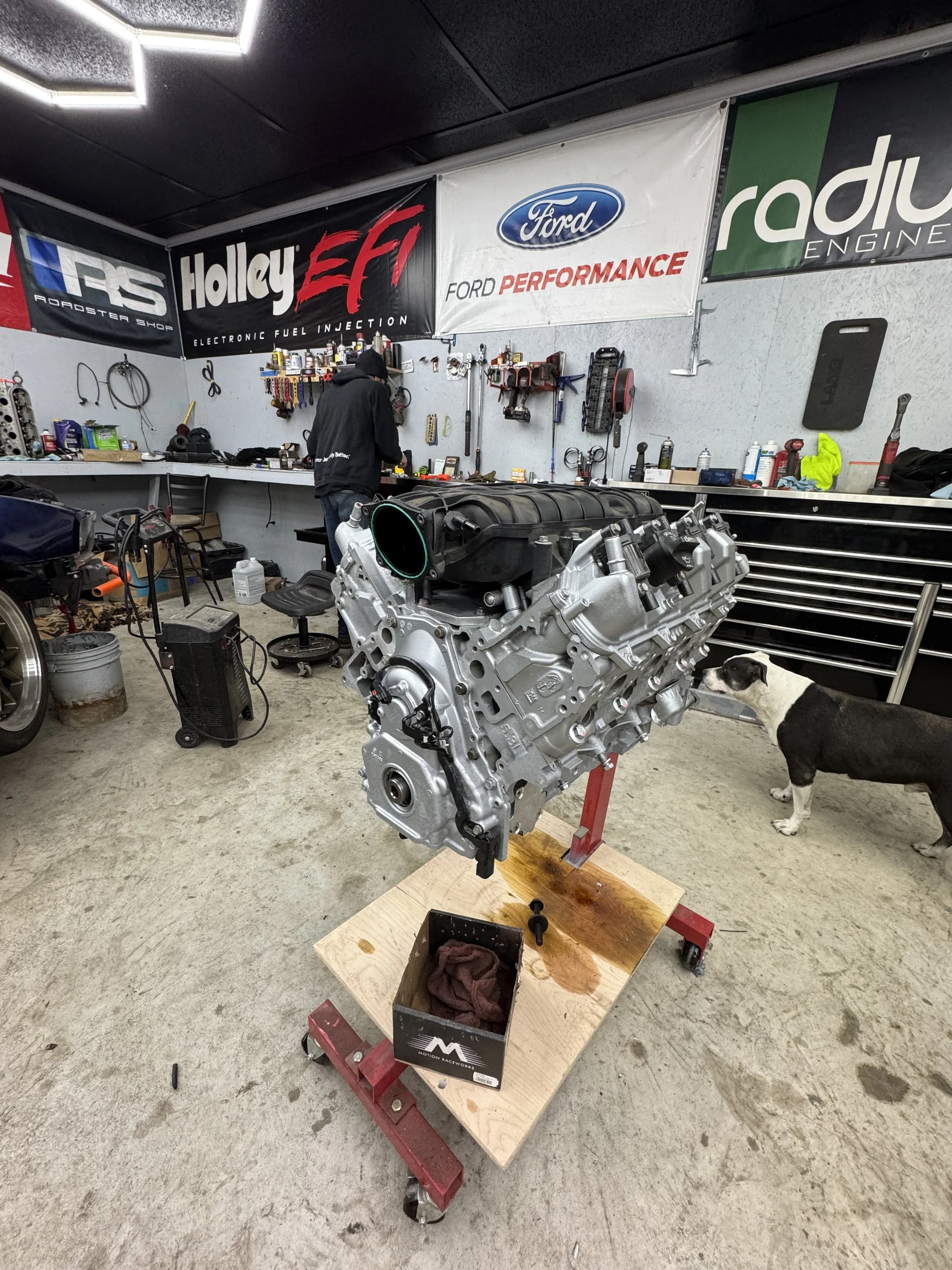 A car engine mounted on a red engine stand in a workshop, with tools and supplies on the workbench behind. The room has banners, including Ford and Holley, on the wall, and a person working at the workbench. A dog standing nearby.