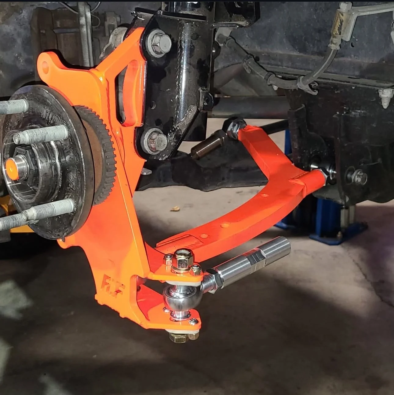 An orange suspension support kit installed on a car's front axle, with the wheel removed, exposing wheel hub, brake components, and suspension parts.