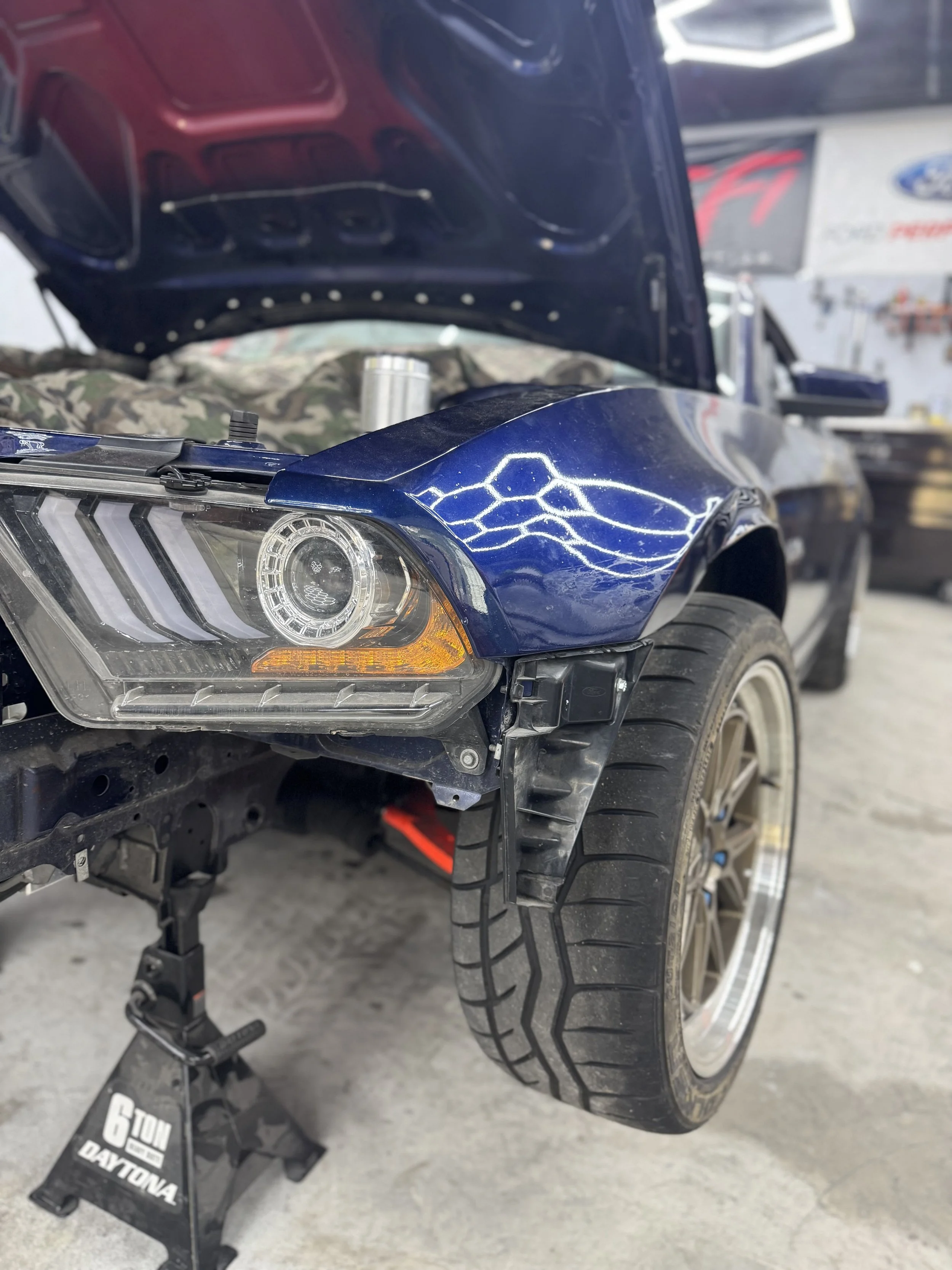 A blue car with its hood open in a garage, showing the front left side, headlight, large tire, and partial engine. The car is on a jack stand labeled '6 TON DAYTONA'.